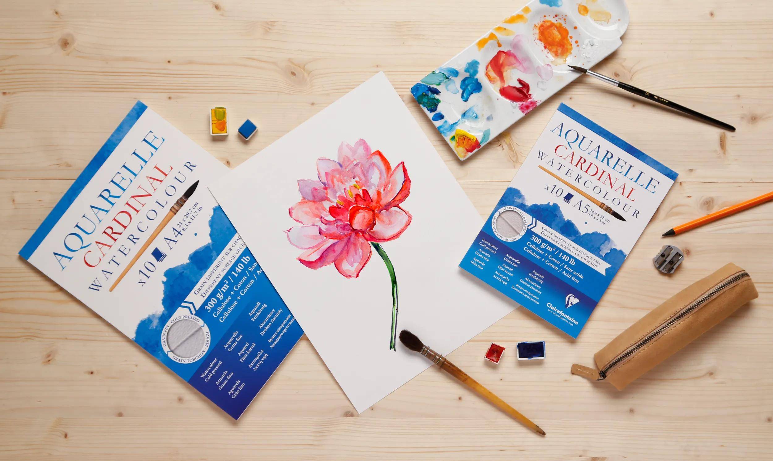 Ein lebendiges rosa Aquarellblumengemälde ist von Malzubehör umgeben, darunter ein Pinsel, eine Palette mit gemischten Farben und Papierverpackung.