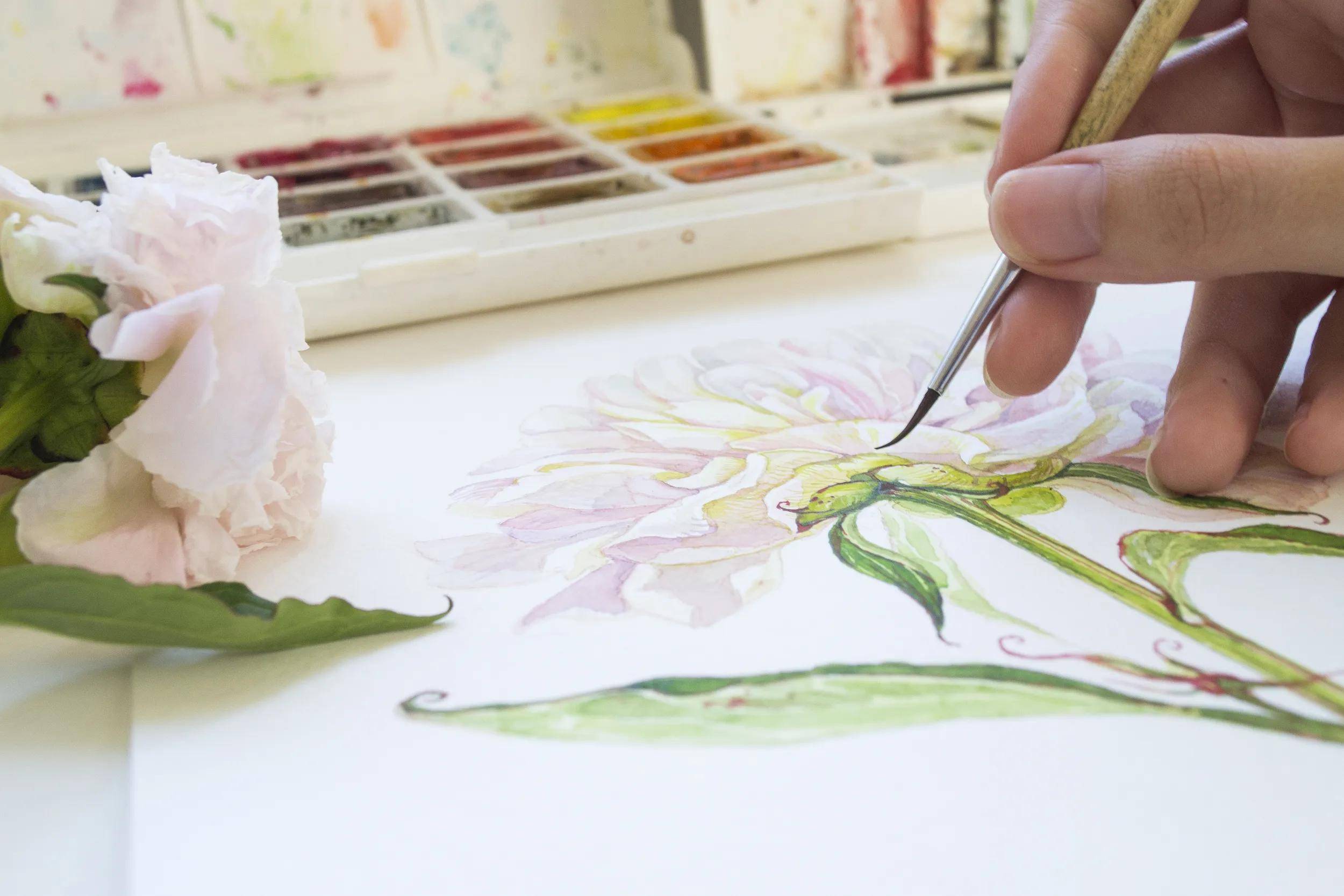 Eine Person malt eine detaillierte Aquarellblume mit grünen Blättern. Ein offenes Farbpalette und eine rosa Blume befinden sich in der Nähe.
