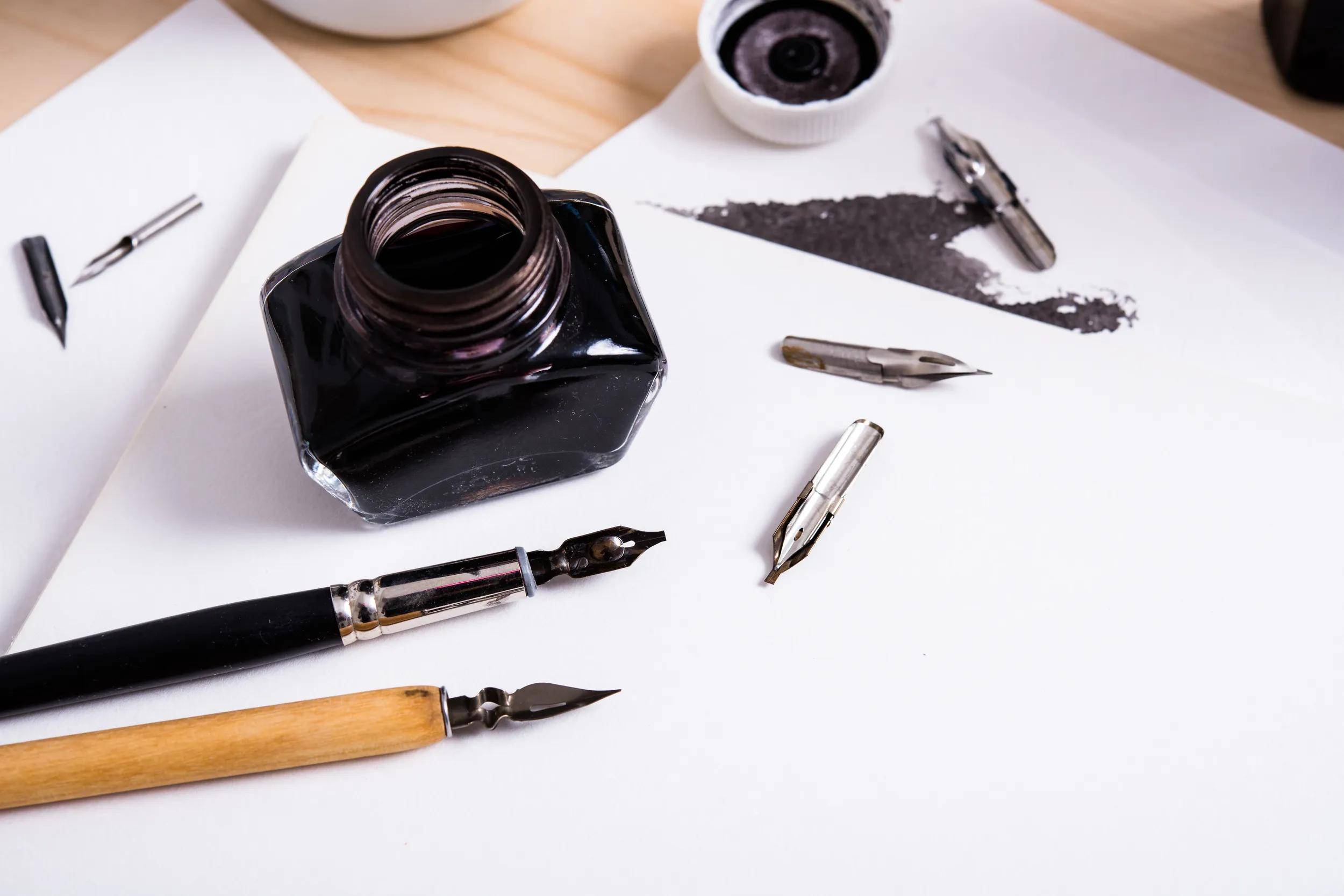 Tintenfass, Füllfederhalter, Federn und ein schwarzer Tintenfleck auf weißem Papier, auf einem Schreibtisch für Kalligraphie oder Handschrift angeordnet.