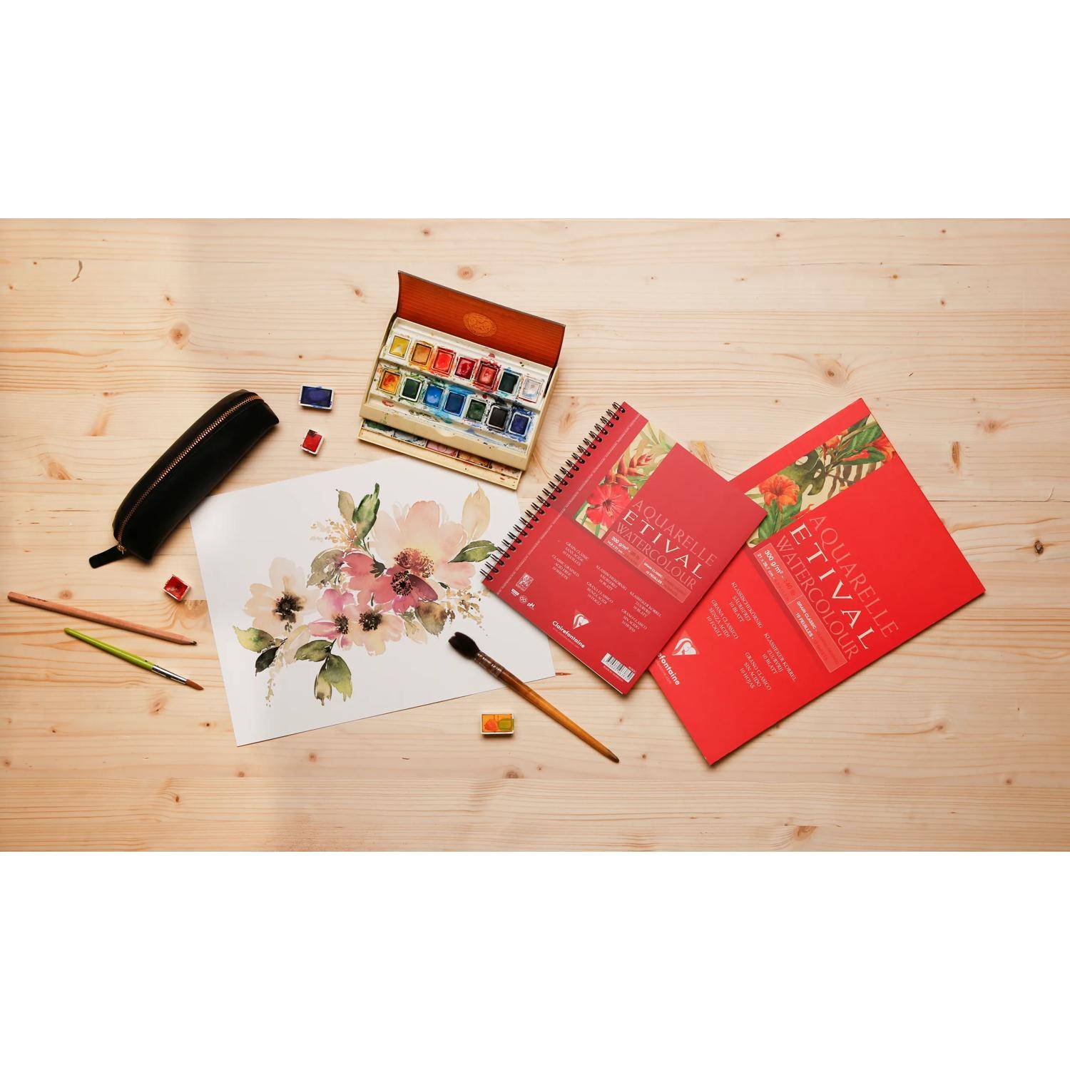 Aquarellmalerei-Zubehör auf einem Holztisch: Malkasten, Pinsel, Skizzenbuch mit Blumenillustration, Stifteetui und rote Notizbücher.