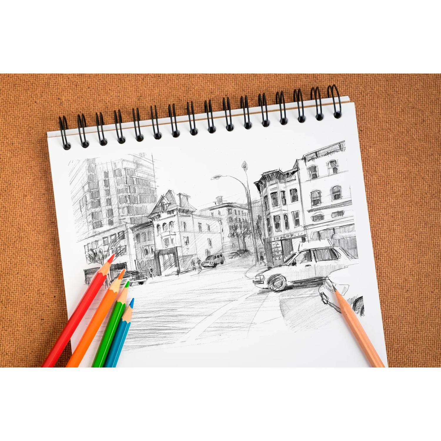 Stifte und ein Skizzenbuch mit einer Bleistiftzeichnung einer Straßenszene einer Stadt, Gebäude, Autos und eine Straßenlaterne auf einer Tischplatte.
