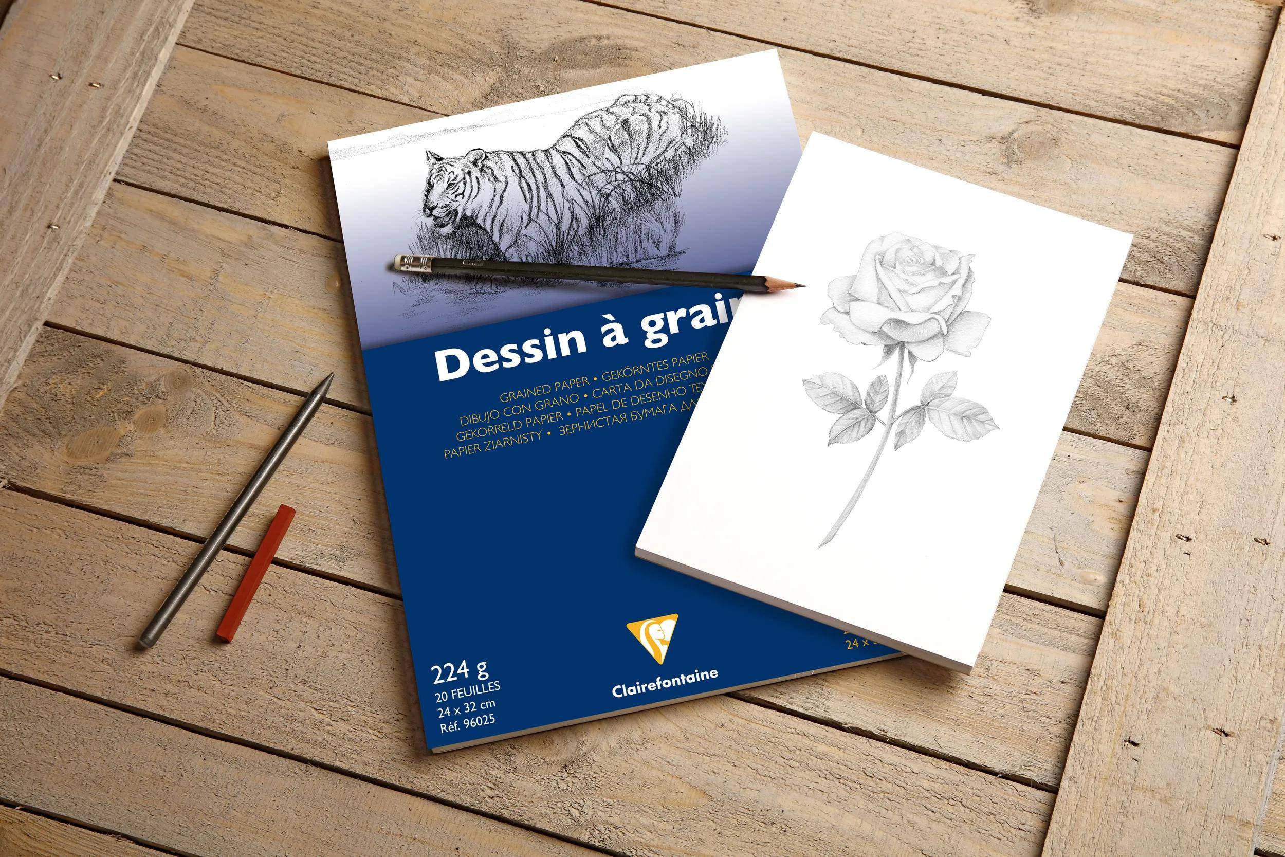 Zeichenmaterialien auf einem Holztisch: ein blaues 'Dessin à gra...'-Papierpad mit einer Tigerzeichnung, ein Bleistift und ein kleines Skizzenbuch mit einer Rosenzeichnung.