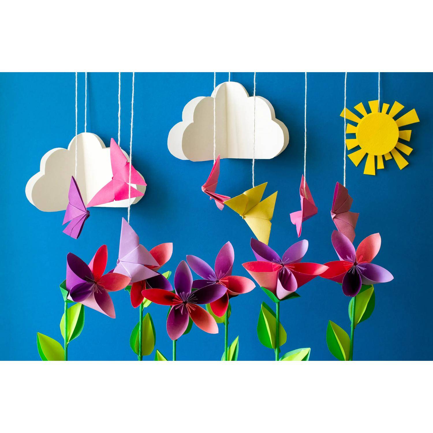 Schmetterlinge und Blumen aus Papier hängen an Schnüren vor einem blauen Himmelshintergrund mit ausgeschnittenen Wolken und einer Sonne.