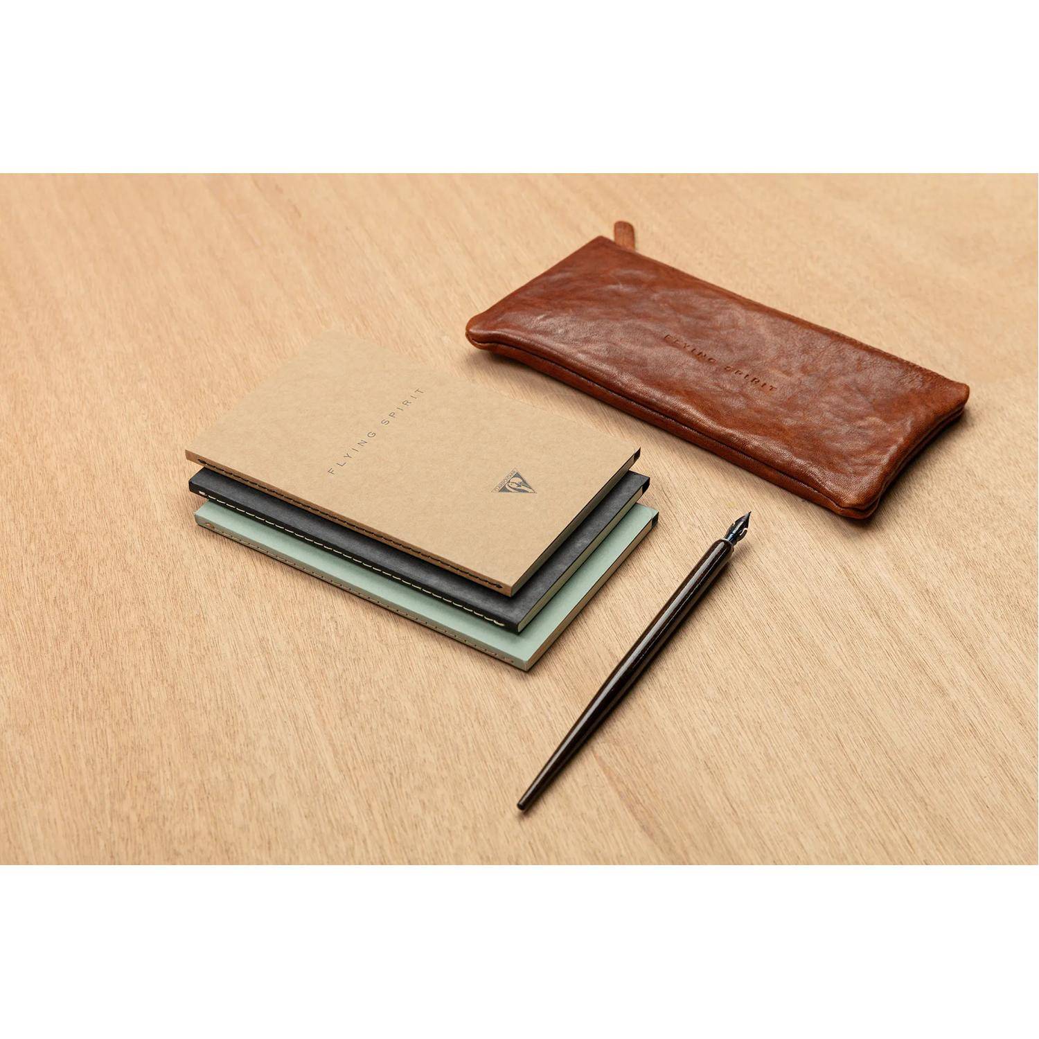 Stapel von Notizbüchern mit einem schwarzen Stift und braunem Leder-Stifteetui auf einem hellholzernen Schreibtisch.