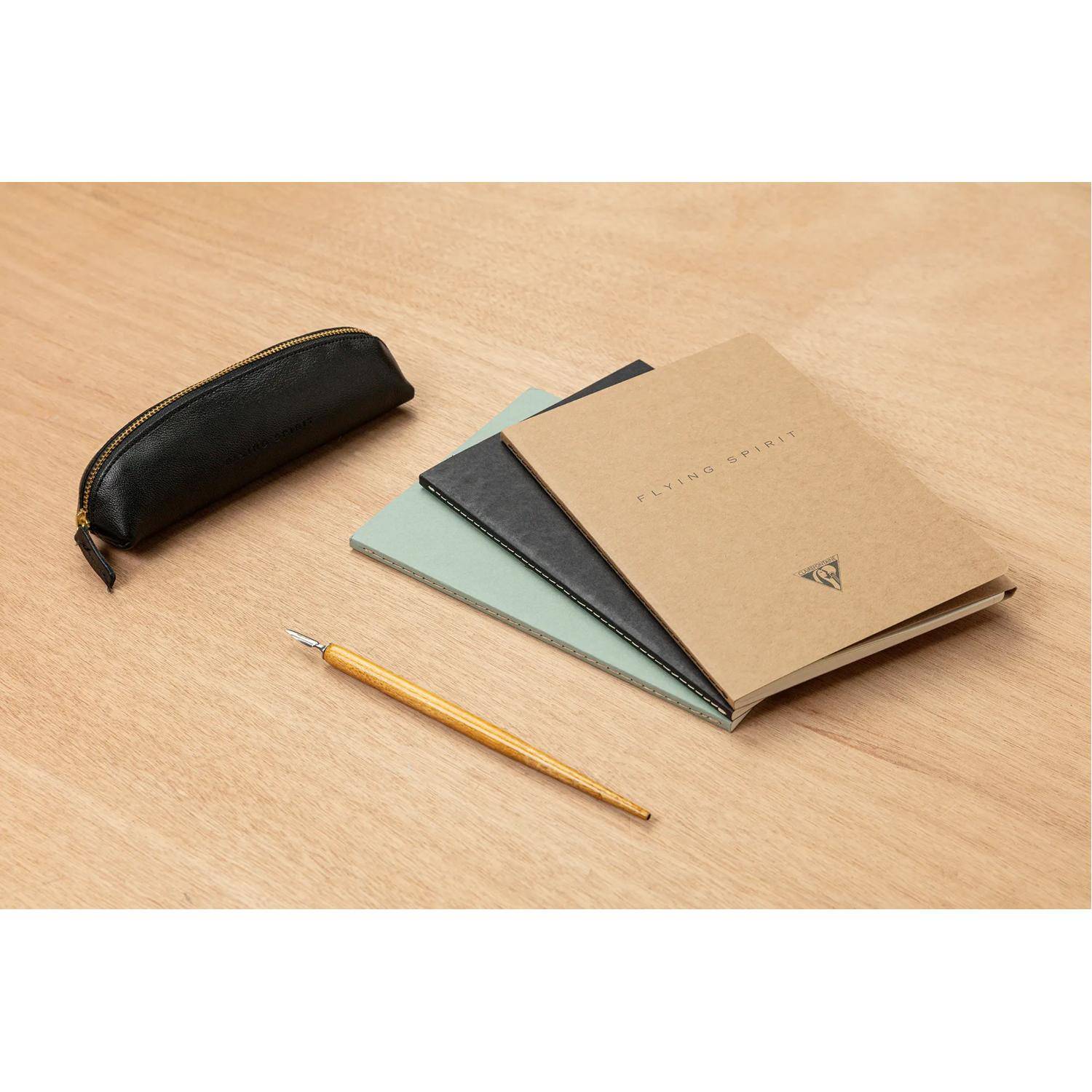Bleistift, schwarzes Reißverschlussetui und gestapelte Notizbücher auf einem Holzschreibtisch, die ein minimalistisches Schreibwarenset zeigen.