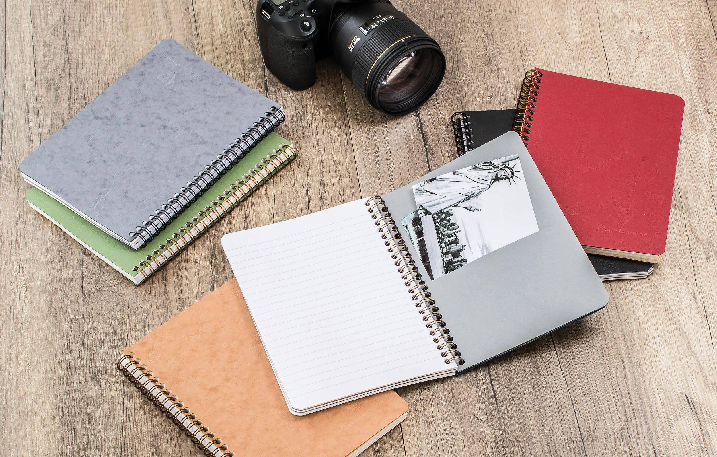 Übersichtsaufnahme von mehreren Spiralnotizbüchern und einer Kamera auf einem Holztisch, mit einem offenen leeren Notizbuch und einem Umschlag, der ein skizzenhaftes Foto zeigt.