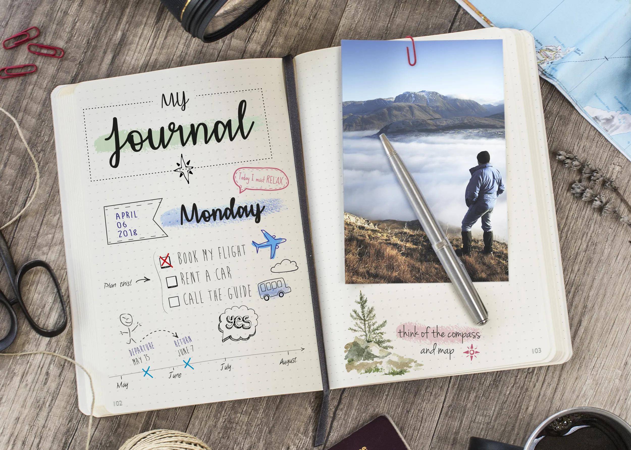 Offenes Journal mit einer Wochenplanseite für Montag und einem eingeklebten Bergfoto, das auf einem Holzschreibtisch mit Stiften und Brillen liegt.