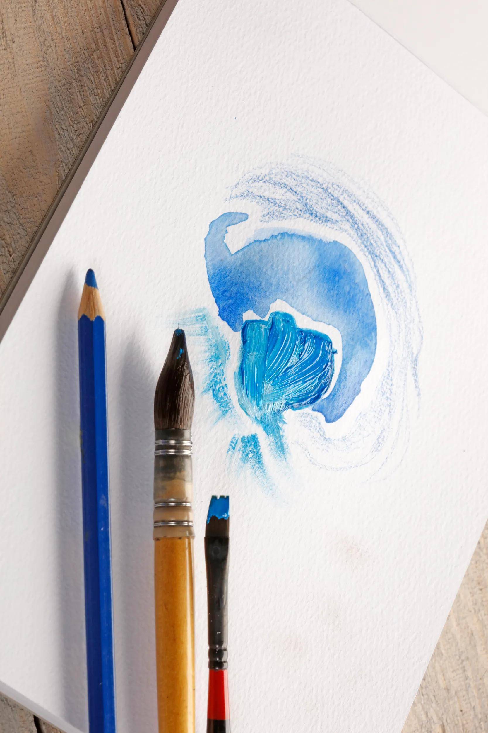 Kunstmaterialien auf Papier: ein blauer Stift und zwei Pinsel neben einem kleinen abstrakten blauen Aquarellgemälde.