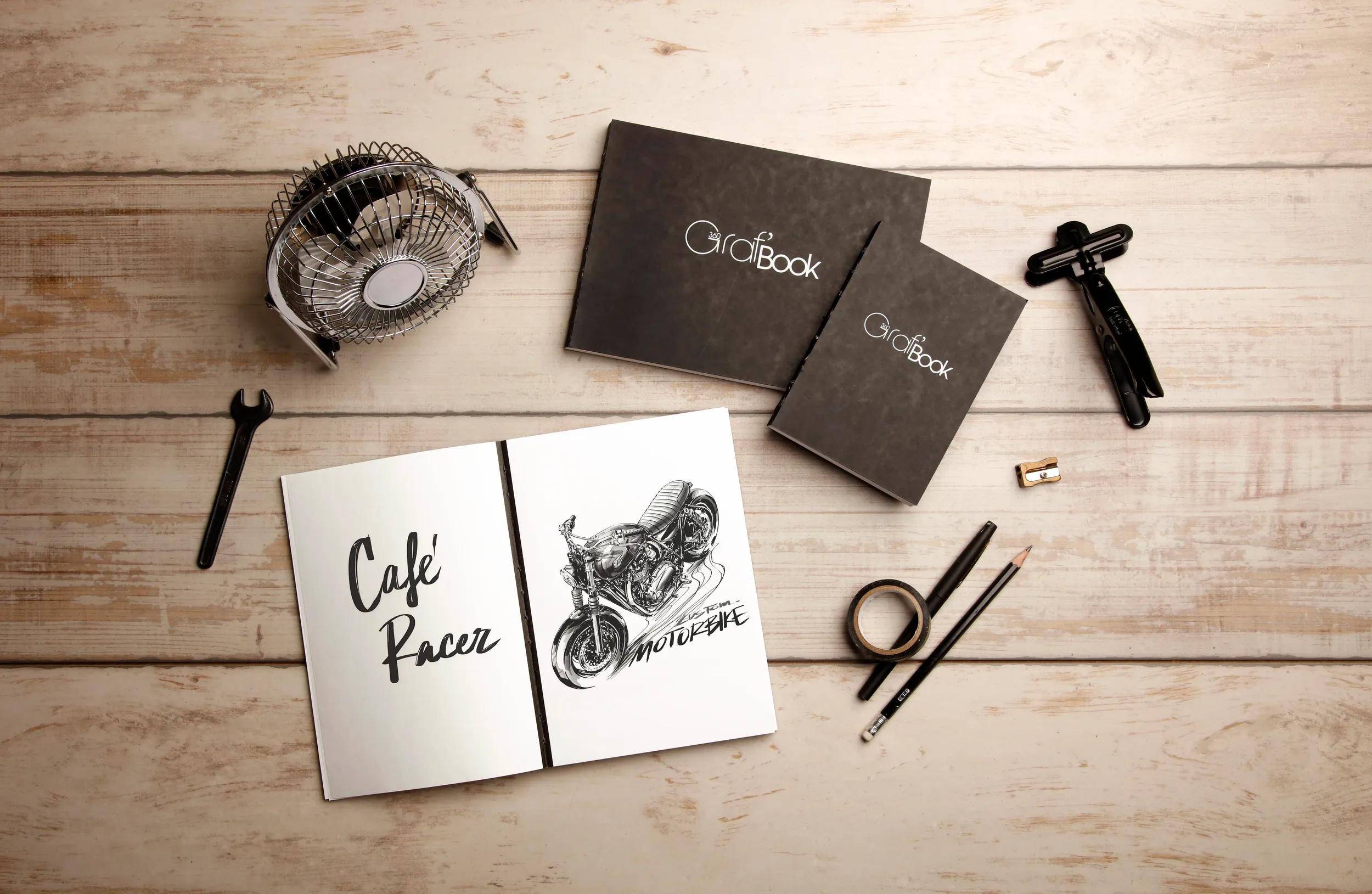 Draufsicht auf ein im Café-Racer-Stil gestaltetes Skizzenbuch, das auf einer Motorradzeichnung aufgeschlagen ist und von Notizbüchern und Kunstwerkzeugen auf einem Holzschreibtisch umgeben ist.