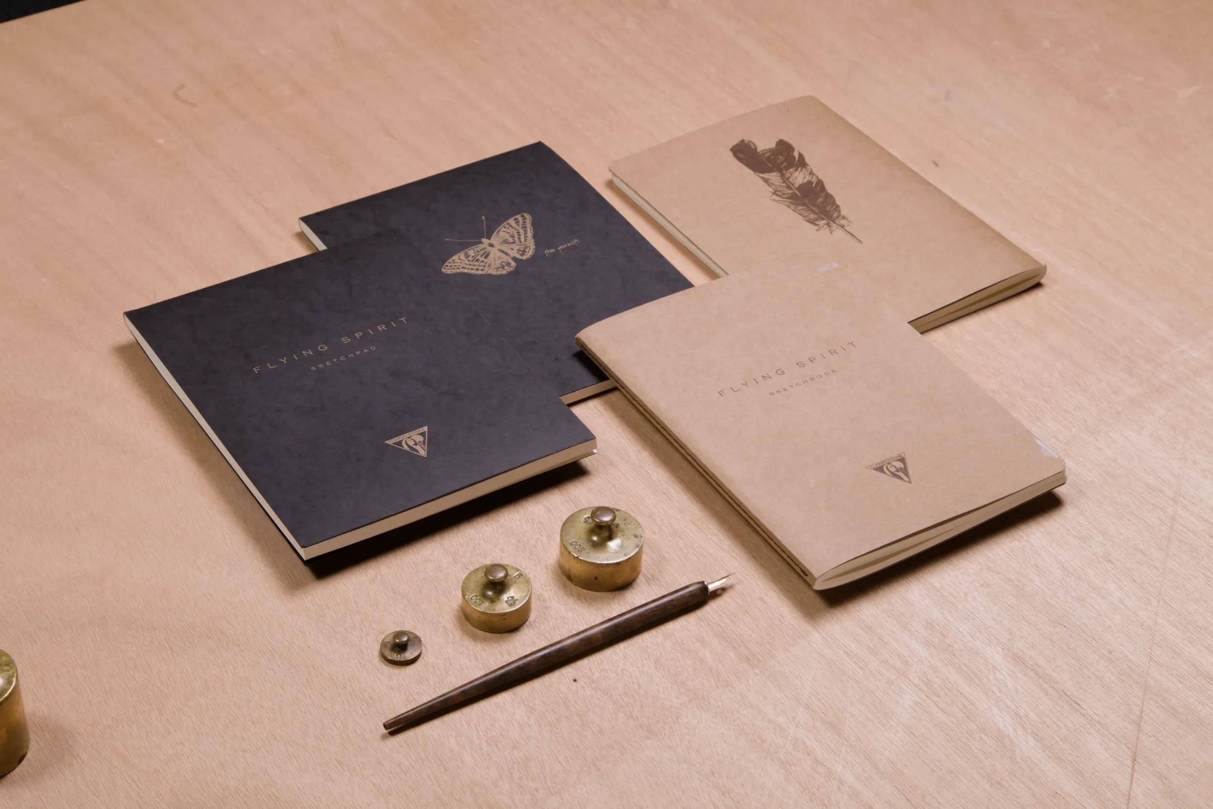 Vier kleine Broschüren oder Notizbücher mit minimalistischen Schmetterlings- und Insektenumschlägen liegen auf einem Holztisch, neben Metallwerkzeugen und Beschlägen.