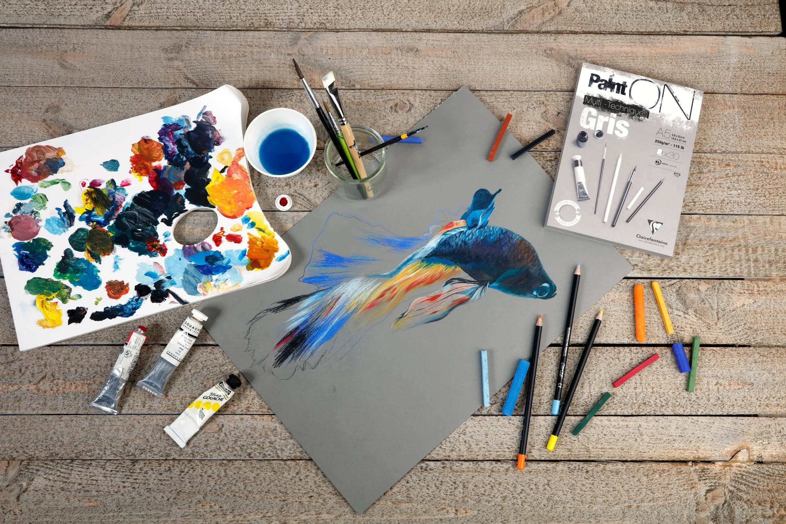 Kunstbedarf arrangiert um eine graue Zeichnung herum, die einen bunten Fisch zeigt, mit Farben, Pinseln, Bleistiften und einem Malbuch auf einer Holzoberfläche.