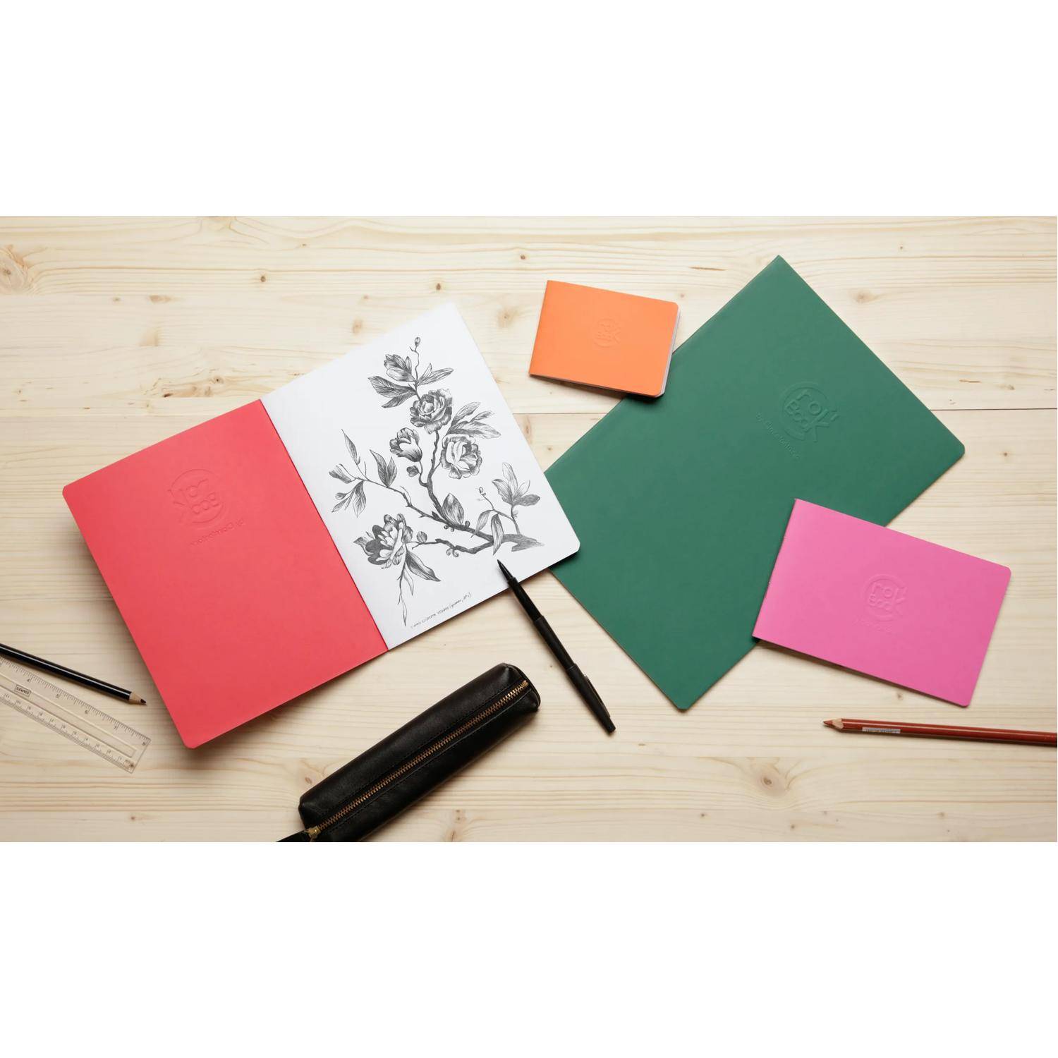 Kunstmaterialien auf einem Holzschreibtisch mit einem offenen Skizzenbuch, das eine Blumenzeichnung zeigt, umgeben von farbigen Notizbüchern, Bleistiften, einem Lineal und einem Radiergummi.