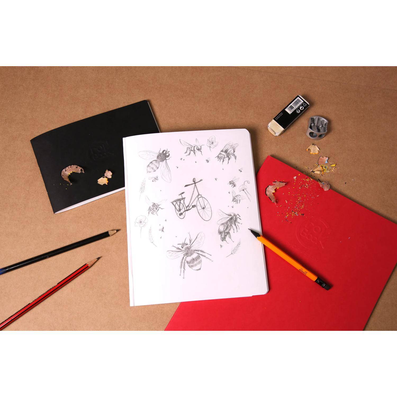 Draufsicht eines Skizzenbuchs mit Insektenzeichnungen und einer Fahrradskizze, umgeben von Bleistiften, Radiergummis und farbigem Papier auf einem braunen Schreibtisch.