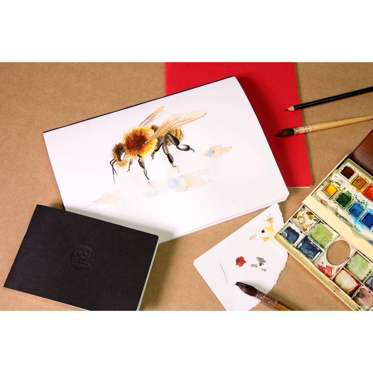 Aquarellmalerei einer Biene auf weißem Papier, umgeben von Pinseln, Palette, Skizzenbuch und Kunstmaterialien auf einer Tischplatte.