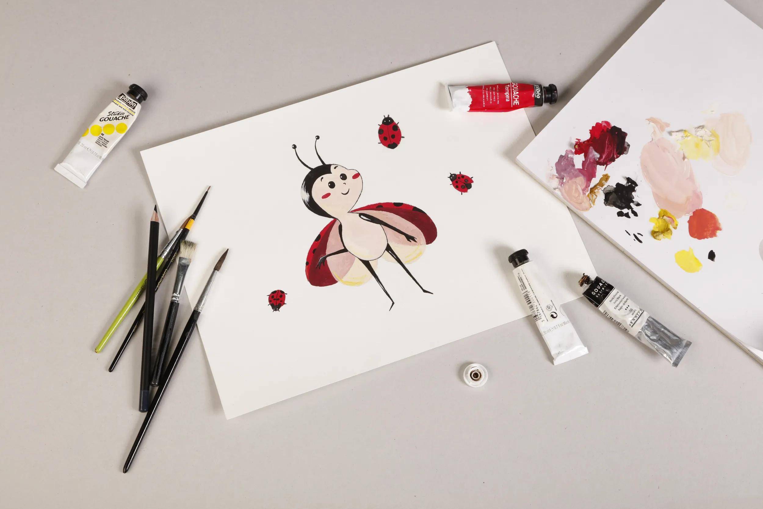 Vogelperspektive eines Aquarellmalerei-Arbeitsplatzes mit einer fertiggestellten Marienkäfer-Illustration auf Papier, Farbtuben, Pinseln, Palette und Farbmustern