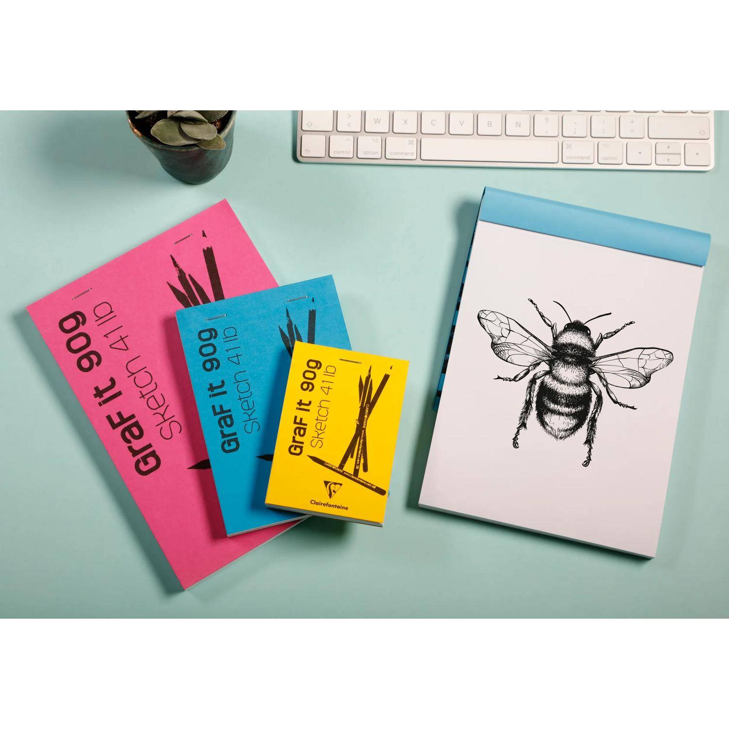Überblick von farbigen Designbüchern und einem Notizbuch mit einer Bienenillustration auf einem Schreibtisch, mit einer teilweise sichtbaren Tastatur und einer Tasse.