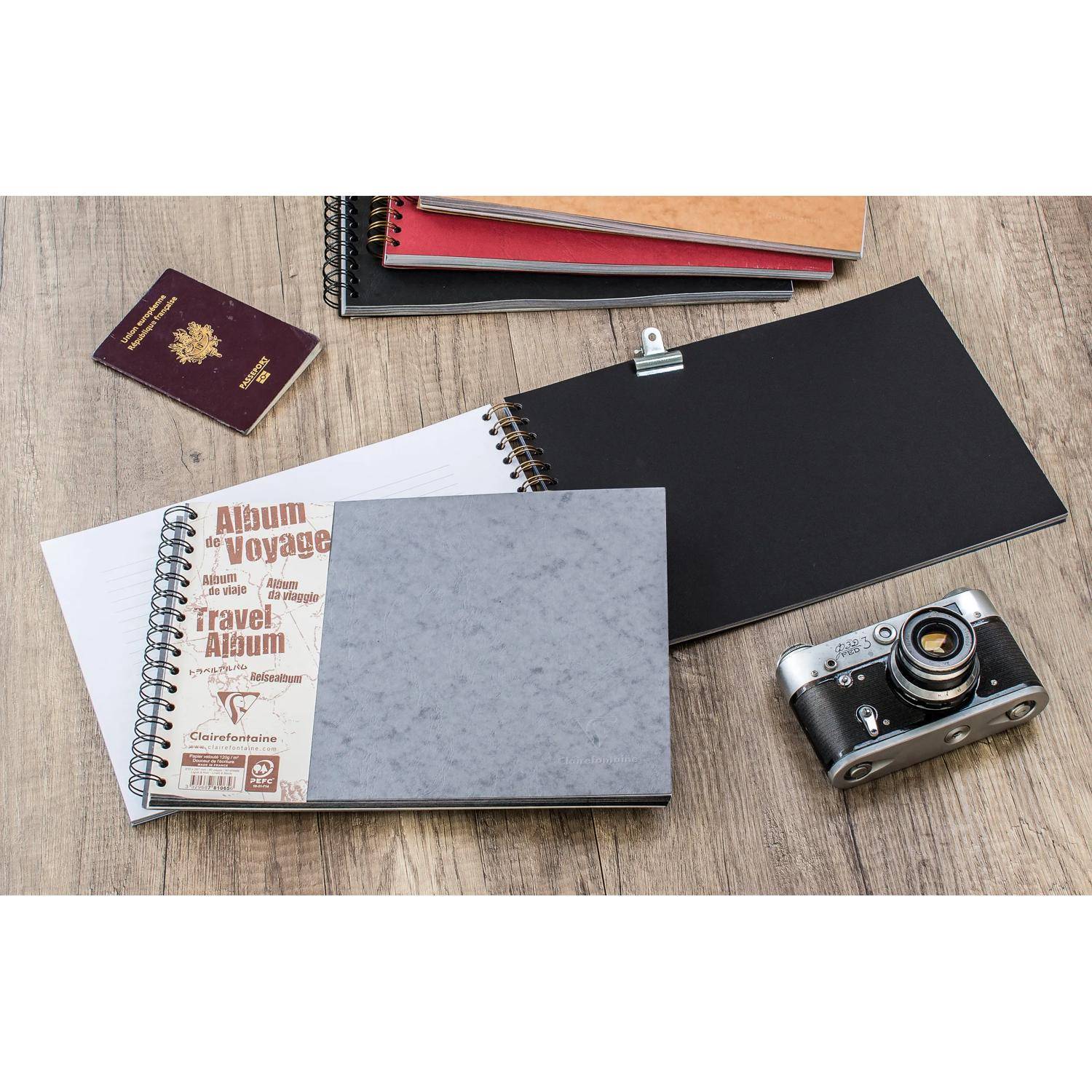 Reisescrapbook-Materialien auf einem Holzboden, darunter mehrere Spiralblöcke, ein Reisepass und eine Vintage-Kamera.