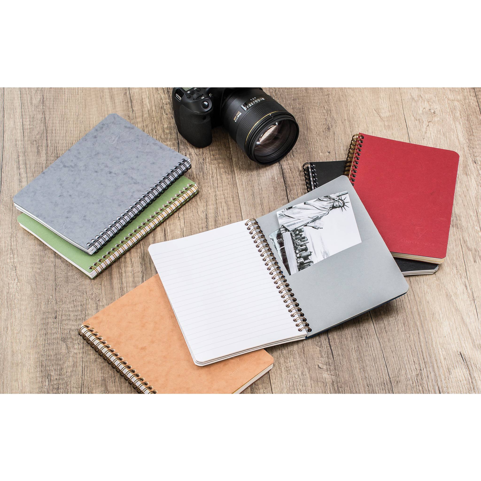 Eine Kamera umgeben von mehreren Spiralnotizbüchern auf einem Holztisch, einschließlich geöffneter und geschlossener Notizbücher in Grau, Rot, Grün, Schwarz und Beige.