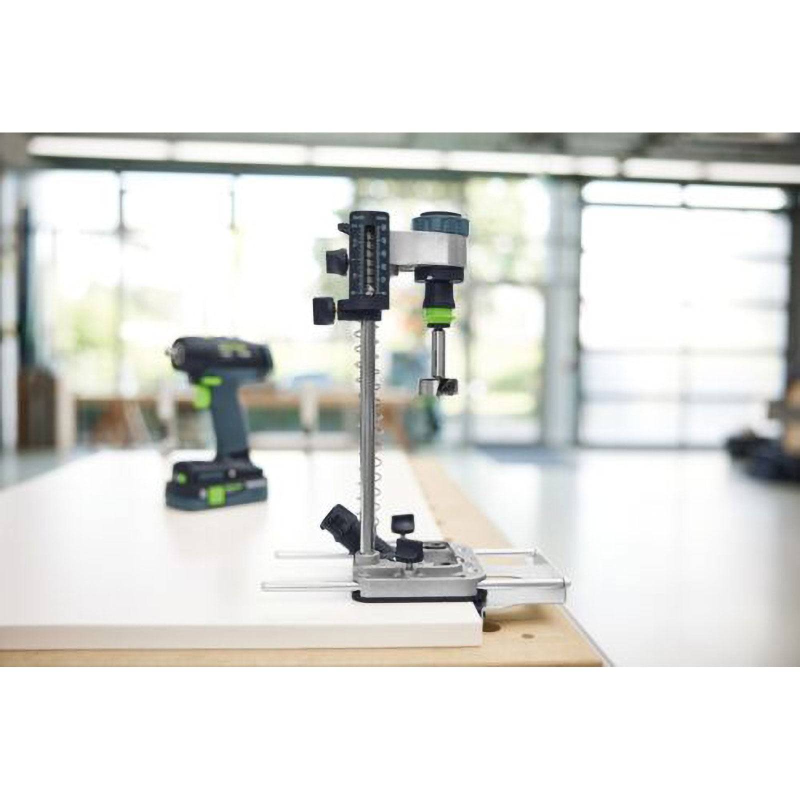 Eine Säulenbohrmaschine steht auf einer Werkbank in einer gut beleuchteten Werkstatt, wobei kabellose Bohrmaschinen im unscharfen Hintergrund zu sehen sind.