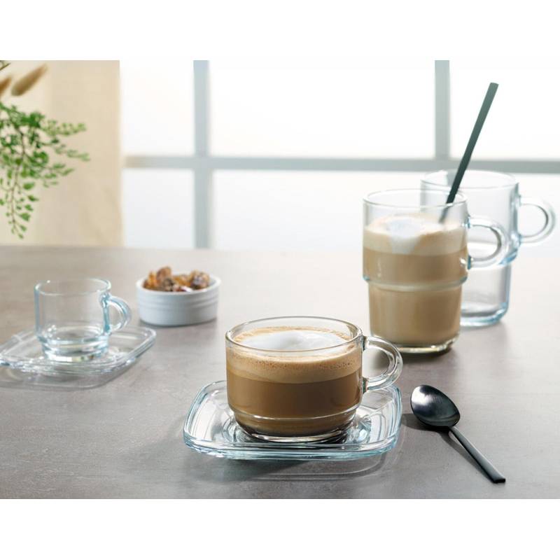 Eine Tasse Kaffee aus Glas steht auf einem Unterteller auf einem Tisch und ist scharf abgebildet, daneben liegt ein Löffel. Im Hintergrund sind zwei weitere Tassen zu sehen.