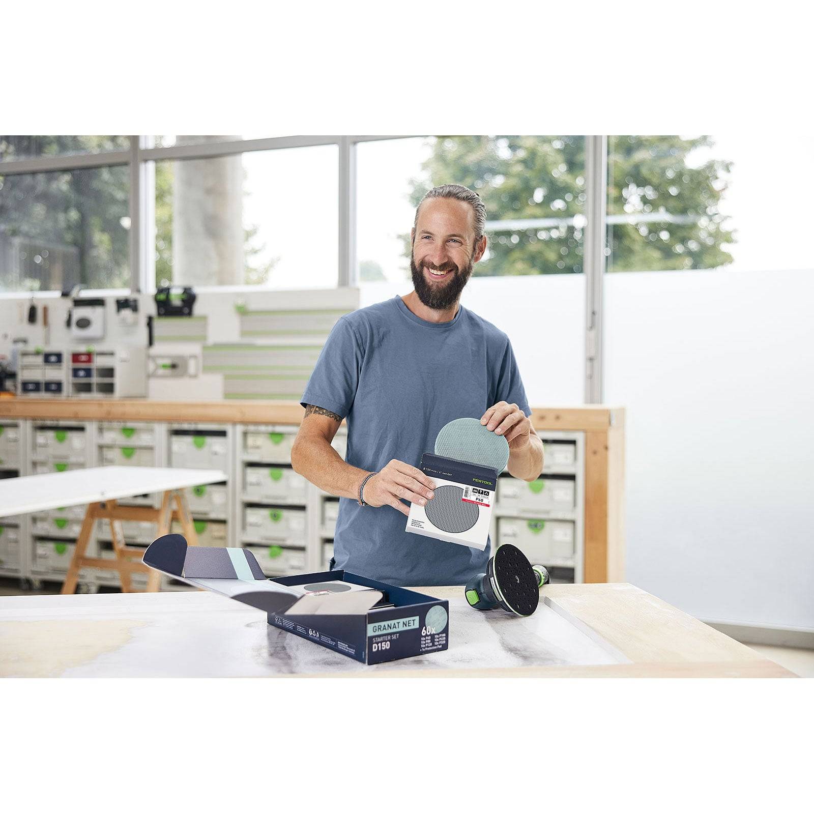 Mann lächelt und steht an einem Tisch mit geöffneten Kartons von Festool-Schleifpapier in einer Werkstatt, wobei er eine positive Arbeitsatmosphäre vermittelt.