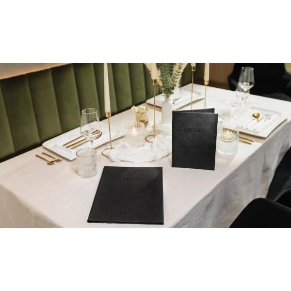 Elegante Restauranttischdeckung mit weißer Tischdecke, goldenem Besteck, Gläsern, Kerzen und schwarzen Menükarten