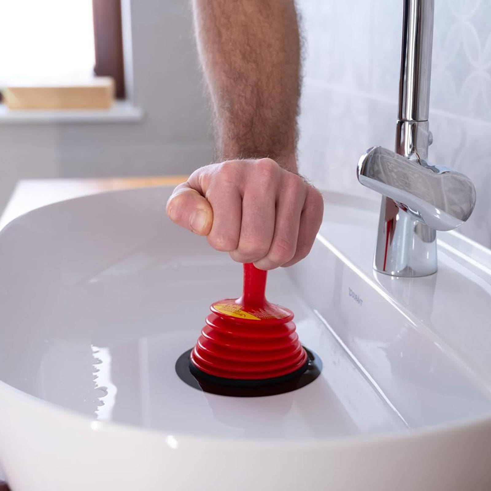 Eine Hand, die einen roten Abflussreiniger (Sauger) verwendet, um ein verstopftes weißes Waschbecken zu entfernen, wobei ein glänzender Wasserhahn sichtbar ist.