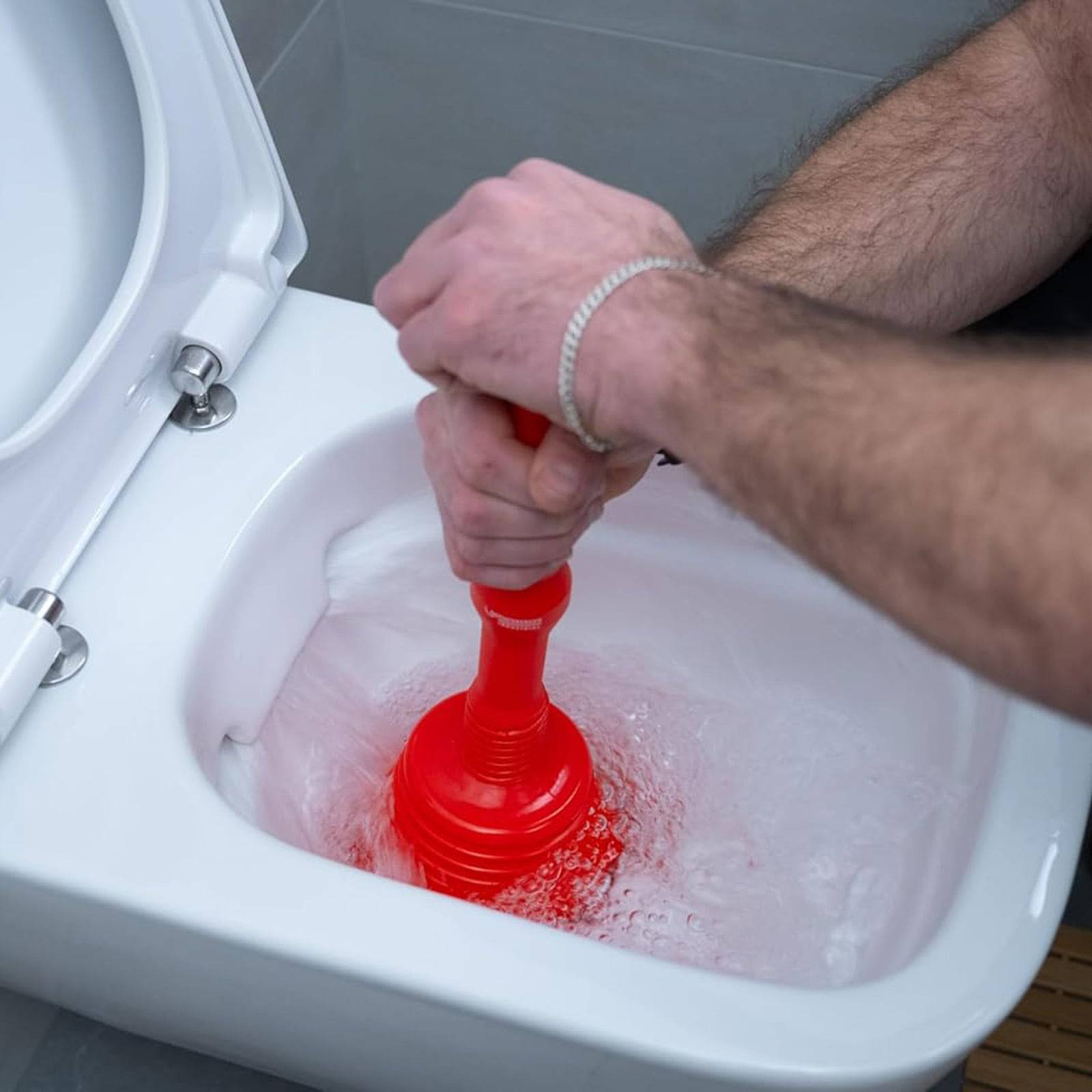 Eine Person benutzt einen roten Kolben in einer weißen, mit Wasser gefüllten Toilette, was darauf hindeutet, dass versucht wird, eine Verstopfung zu beseitigen oder ein Blockierungsproblem zu beheben.