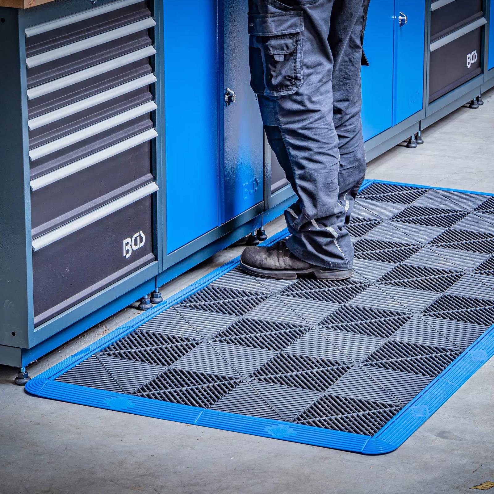 Eine Person in dunkler Hose steht auf einer strukturierten Gummimatte vor blauen Werkzeugschränken, höchstwahrscheinlich in einer Werkstattumgebung.