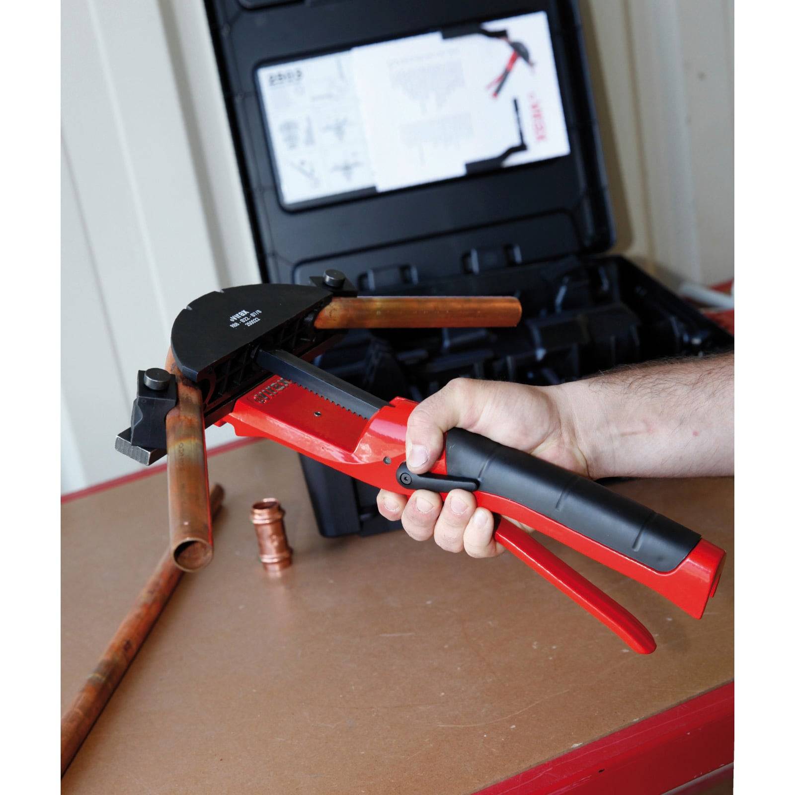 Eine Person benutzt einen roten Rohrbieger, um ein Kupferrohr zu biegen, wobei sich im Hintergrund ein schwarzer Transportkoffer mit Anleitungen und Werkzeugen befindet.