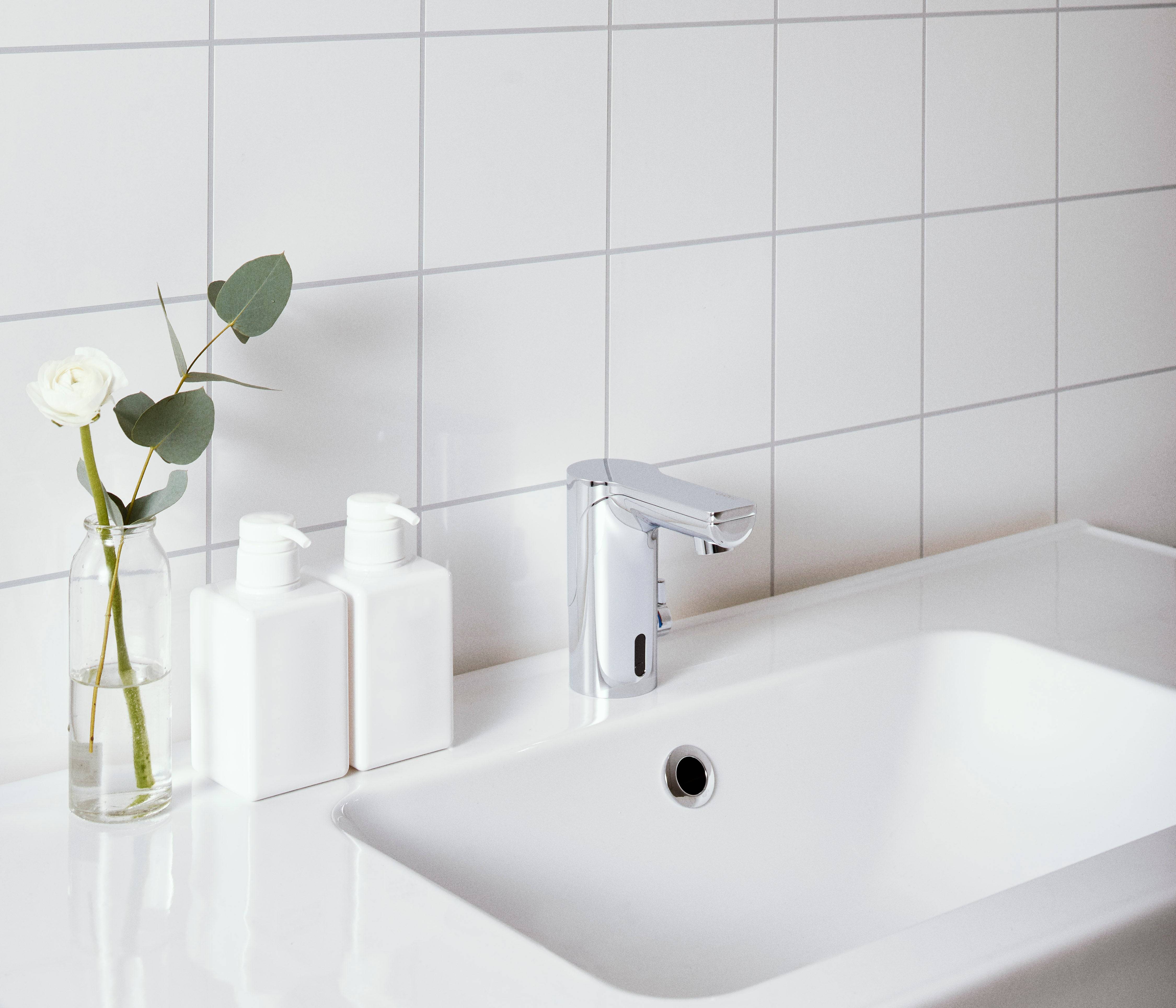 Minimalistisches weißes Badezimmerbecken mit Chromarmatur, zwei weiße Flaschen und eine Vase mit einer weißen Blume auf der Ablage.