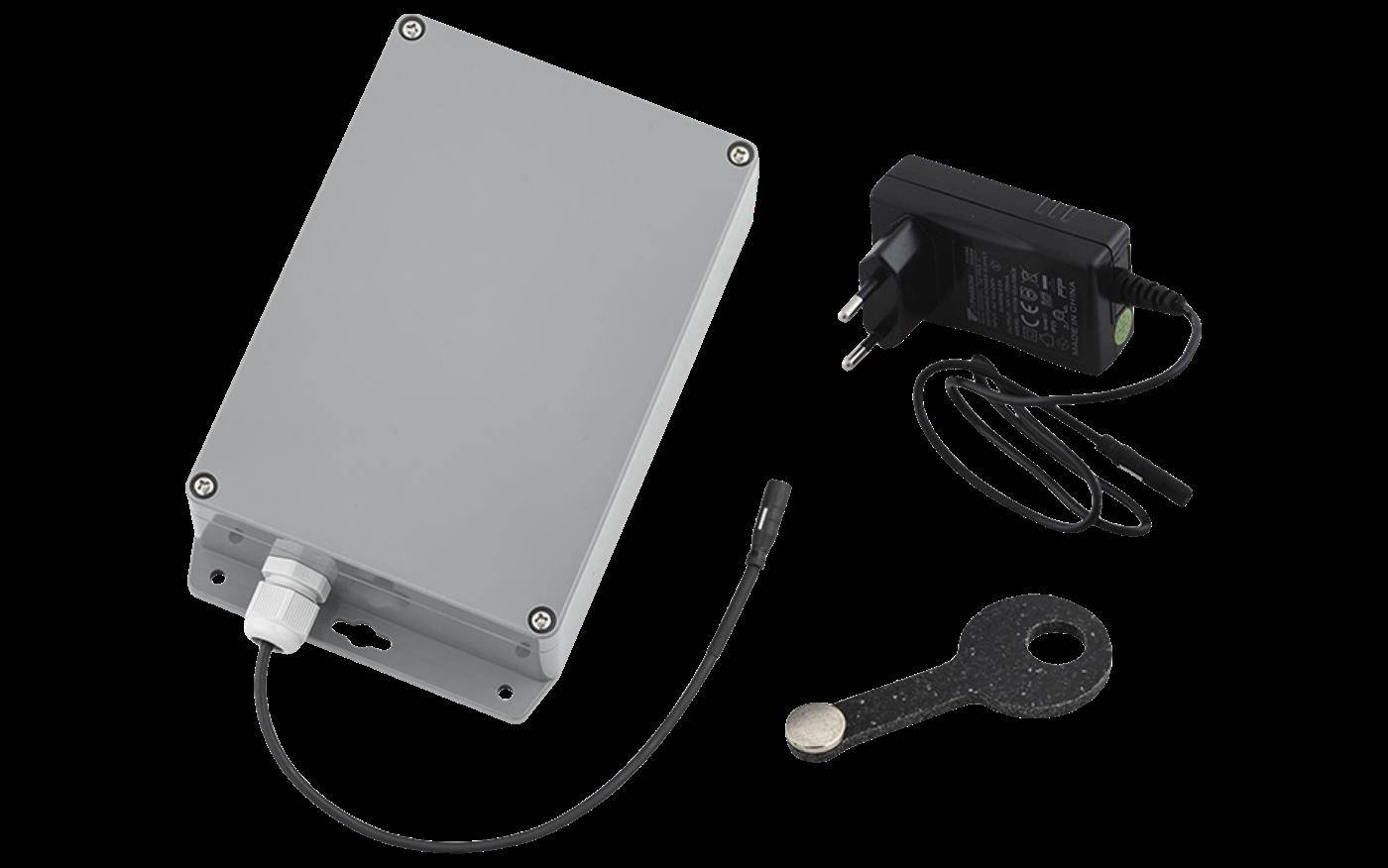 Ein kleines graues Elektronikgehäuse mit angeschlossenem Kabel, einem Stromadapter und einem Montageschlüssel/Werkzeug auf schwarzem Hintergrund.
