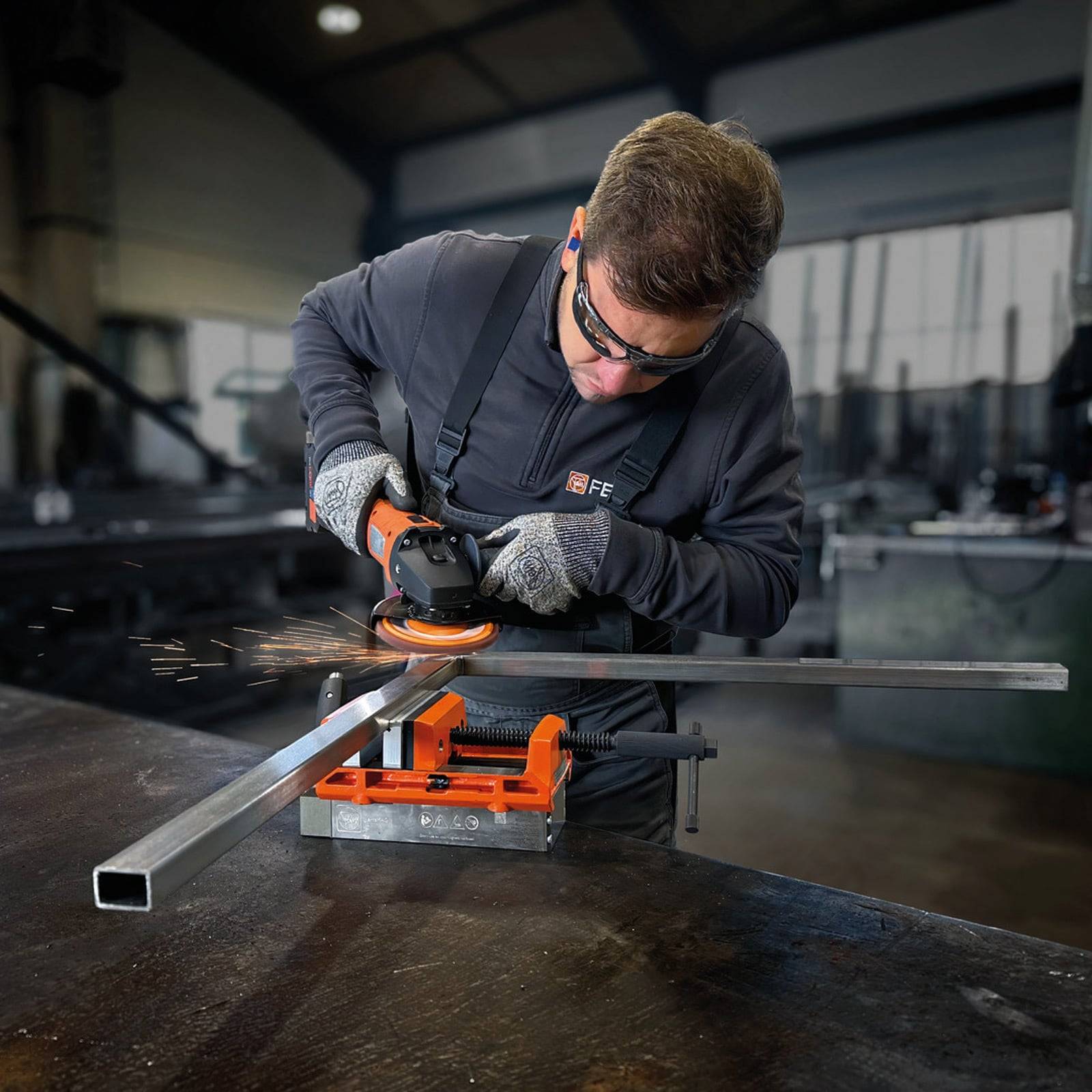 Arbeiter in einer Werkstatt verwendet einen Elektrowinkelschleifer an einem Metallstab, wobei Funken sprühen. Zum Schutz werden Schutzbrille und Gehörschutz getragen.