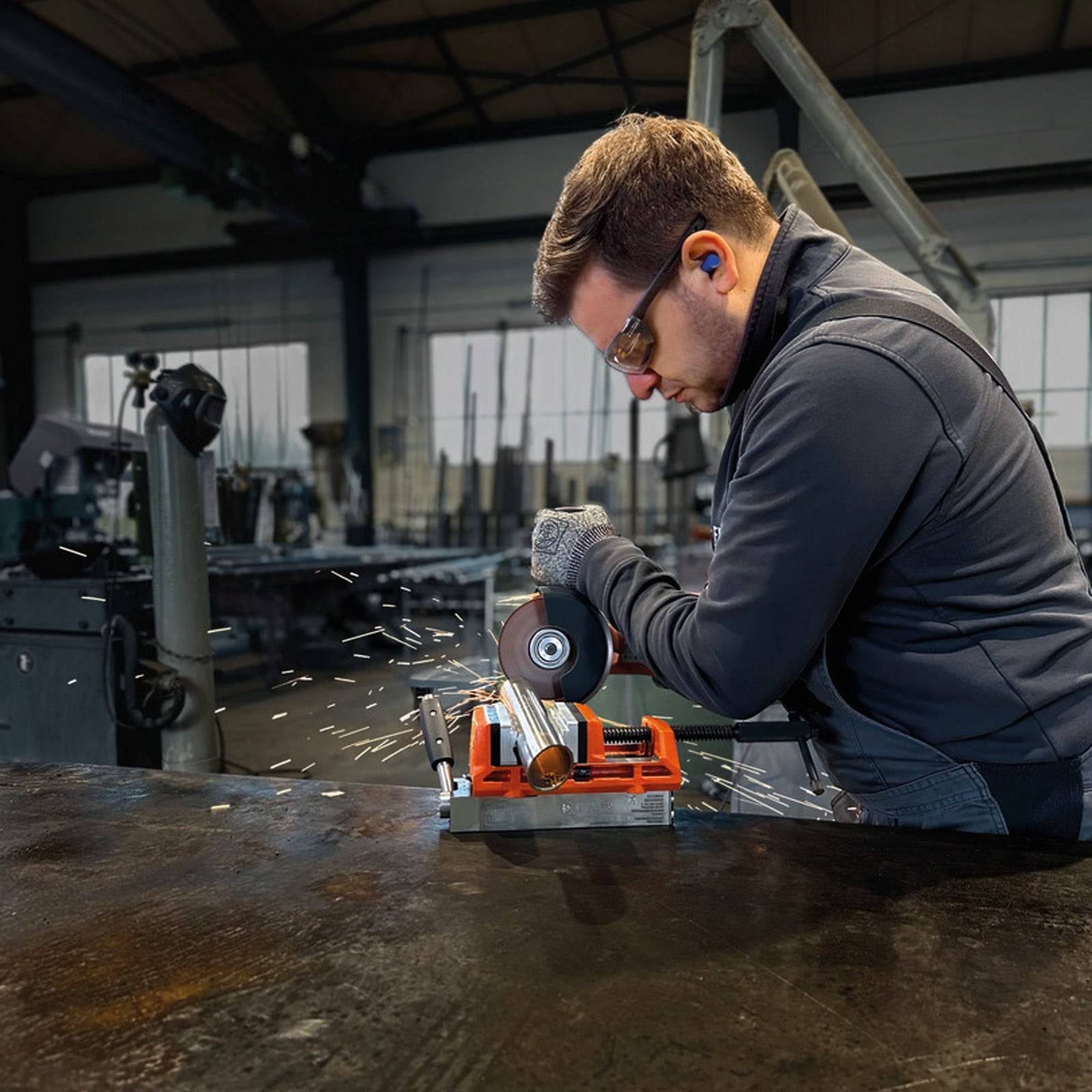 Eine Person in Sicherheitsausrüstung bedient einen Winkelschleifer an einem Metallstück in einer Werkstatt, wobei Funken entstehen. Im Hintergrund sind industrielle Maschinen sichtbar.