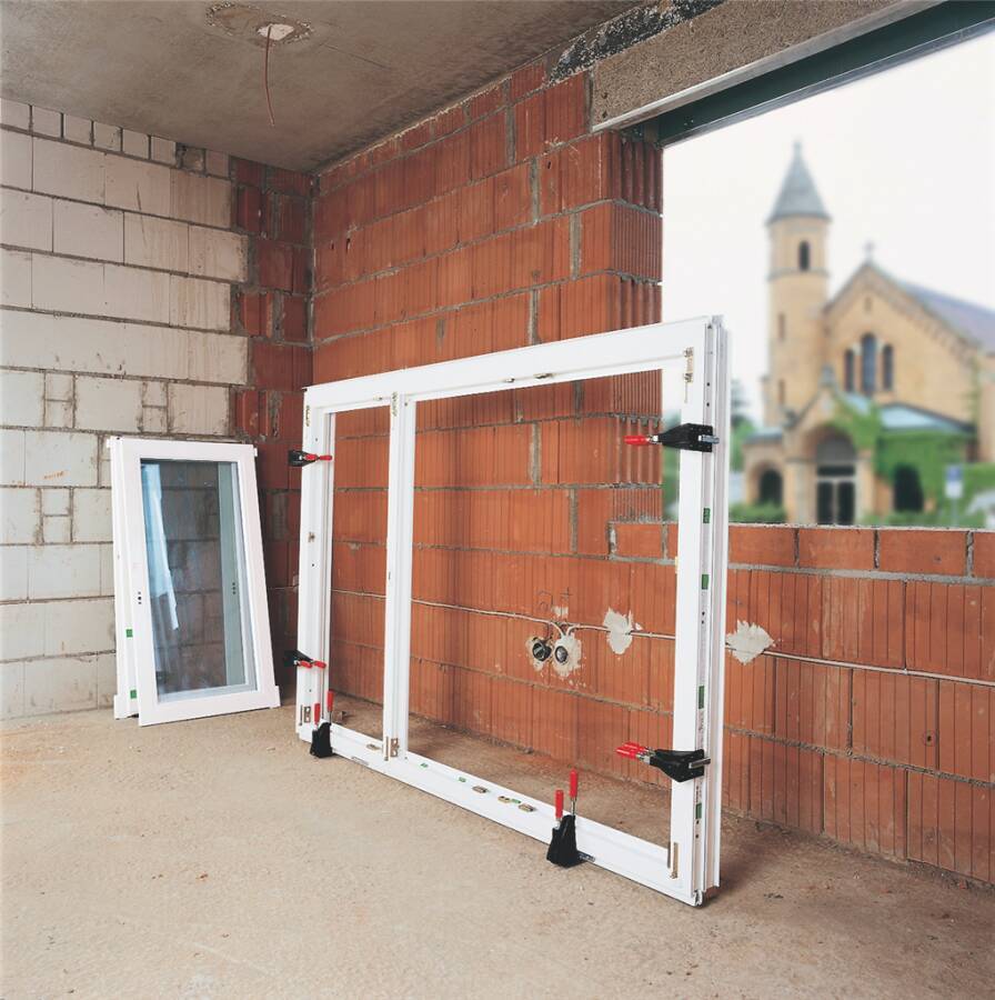 Teilweise installierter weißer Fensterrahmen und separates Glasschiebefenster lehnen an einer unvollendeten Ziegelmauer auf einer Baustelle.
