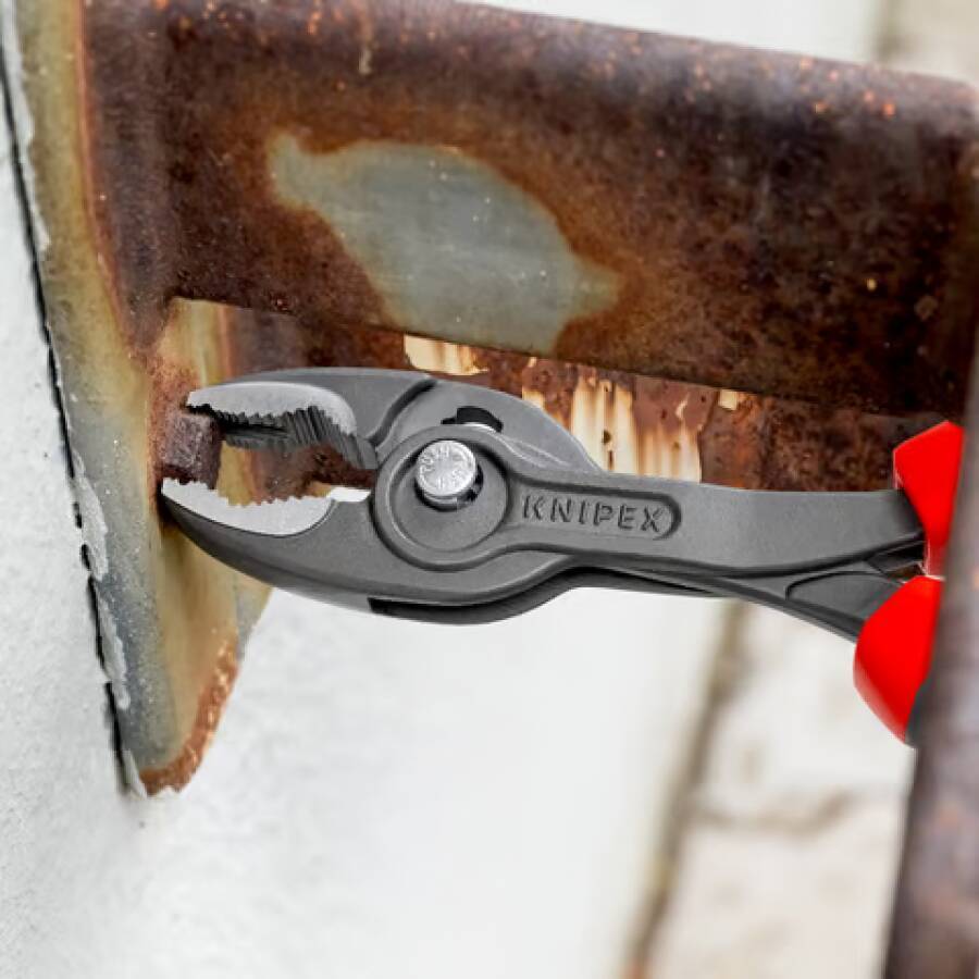 Nahaufnahme von KNIPEX-Zangen, die eine rostige Metallklammer auf einer weißen Oberfläche greifen, zeigt Korrosion und die roten Griffe des Werkzeugs.
