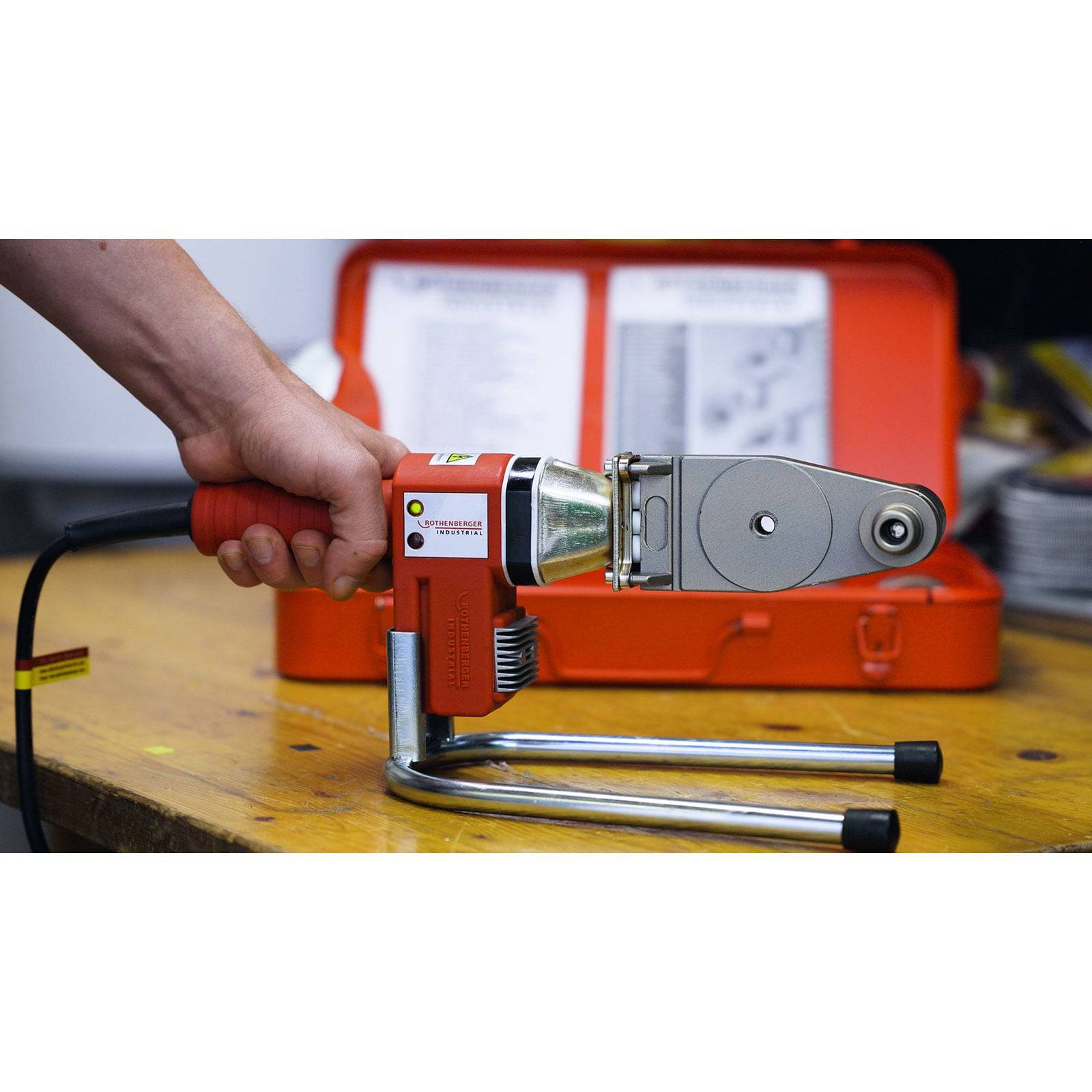 Eine Hand hält ein rotes und silbernes Schweißwerkzeug auf einer Werkbank mit einem orangefarbenen Koffer im Hintergrund, was Einsatzbereitschaft andeutet.