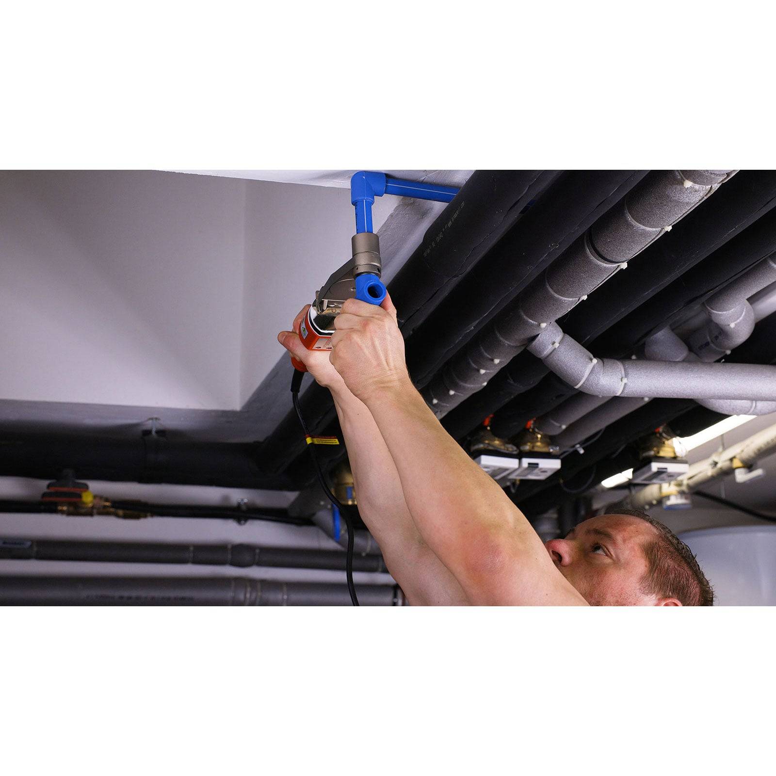 Eine Person arbeitet daran, Rohre an einer Decke zu installieren oder zu reparieren und verwendet dabei ein Werkzeug, um blaue Rohre zwischen schwarzen und grauen Rohren zu justieren.