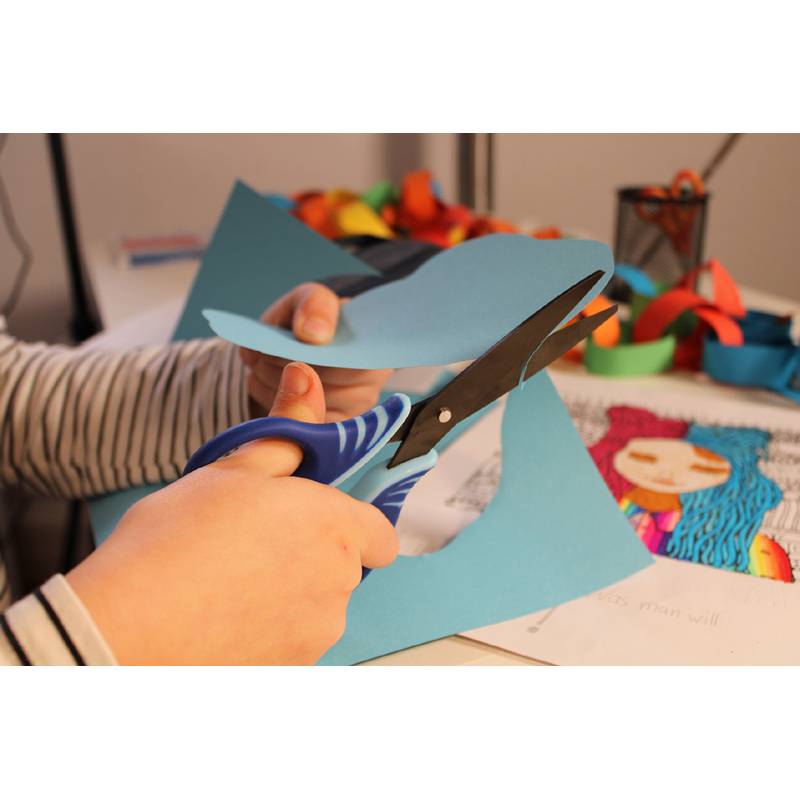 Eine Person schneidet blaues Papier mit einer Schere und sitzt an einem Tisch, der mit bunten Bastelmaterialien gefüllt ist, was auf eine Bastelaktivität hindeutet.