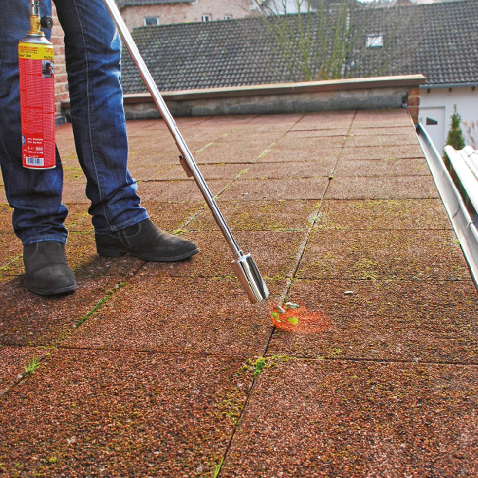 Eine Person verwendet ein langstielige Werkzeug mit einem Gaskanister, um Unkraut zu verbrennen, das zwischen den Fliesen auf einem Dach wächst.