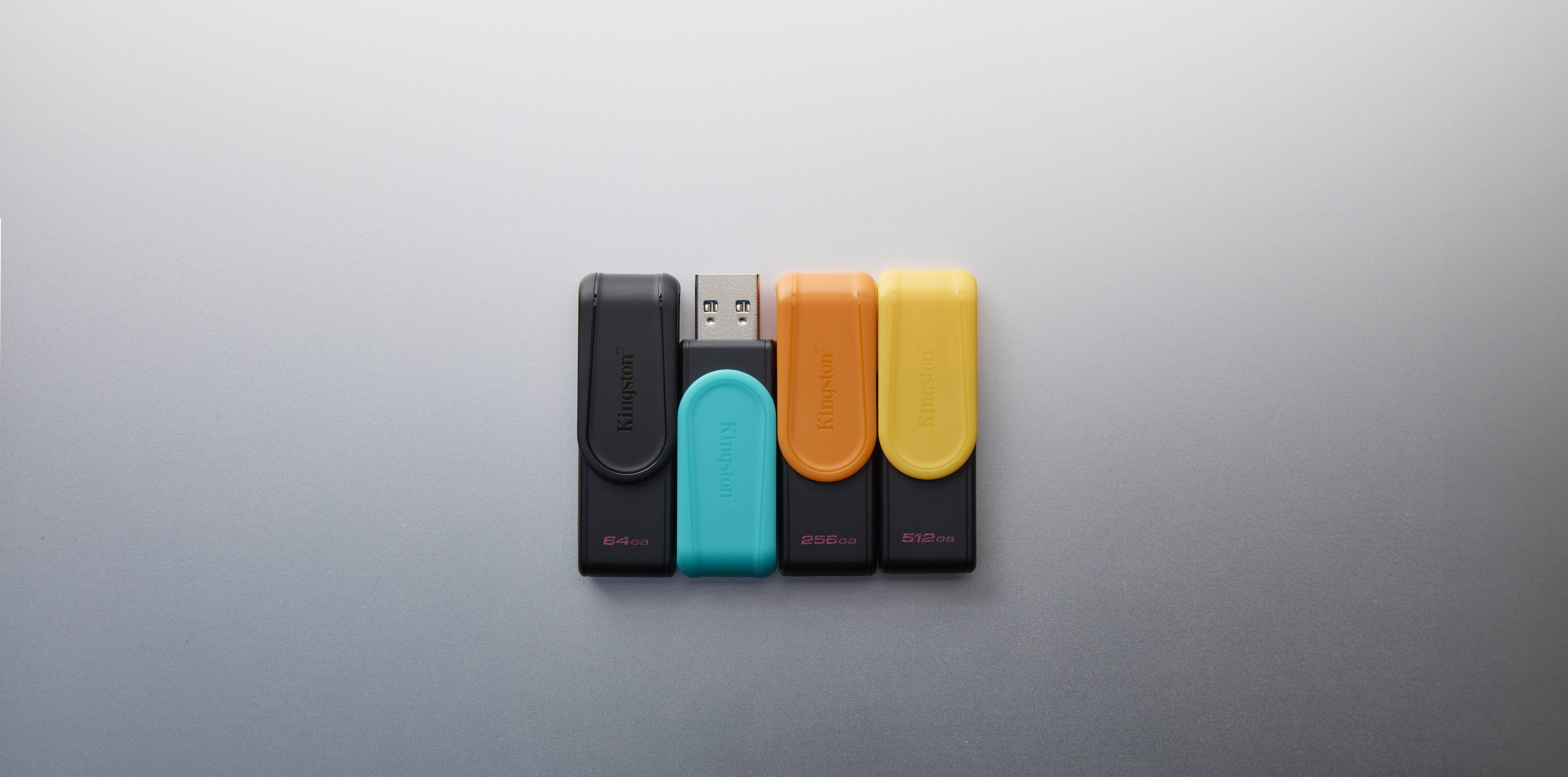 Vier USB-Speichersticks in Schwarz, Cyan, Orange und Gelb, nebeneinander auf grauem Hintergrund angeordnet.