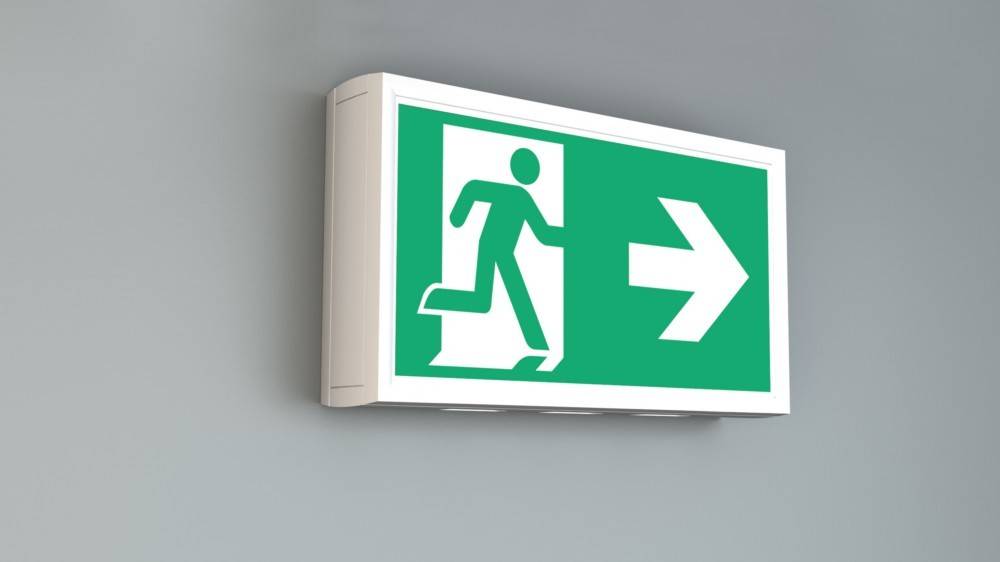 „Notausgangsschild mit einer Figur, die auf eine Tür zulaufend und mit einem nach rechts weisenden Pfeil dargestellt ist, der die Richtung zum nächstgelegenen Ausgang anzeigt.