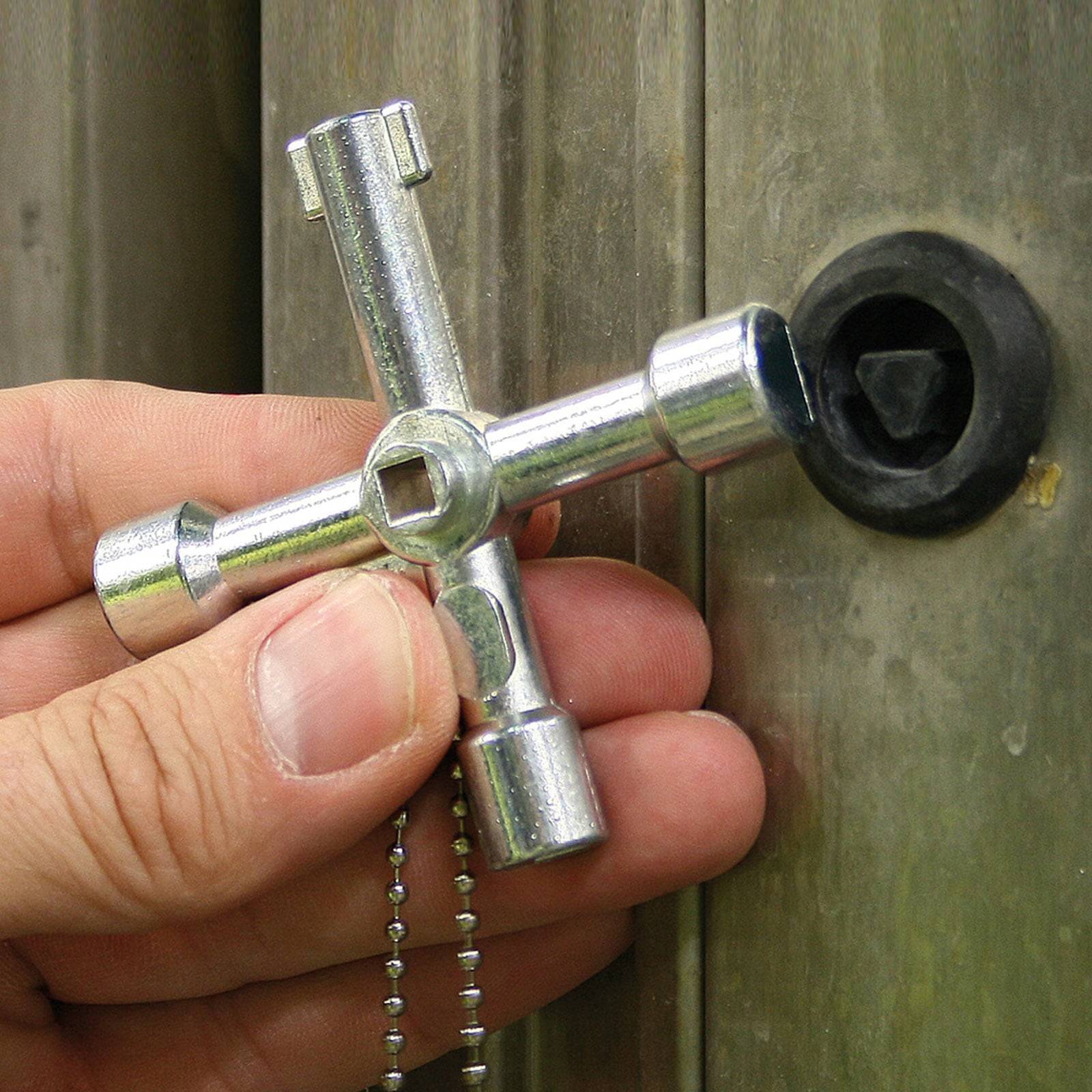 Eine Hand hält einen silbernen Metallutilitätsschlüssel, kreuzförmig geformt, in der Nähe eines kreisrunden Schlosses auf einer Holz- oder Metalloberfläche.
