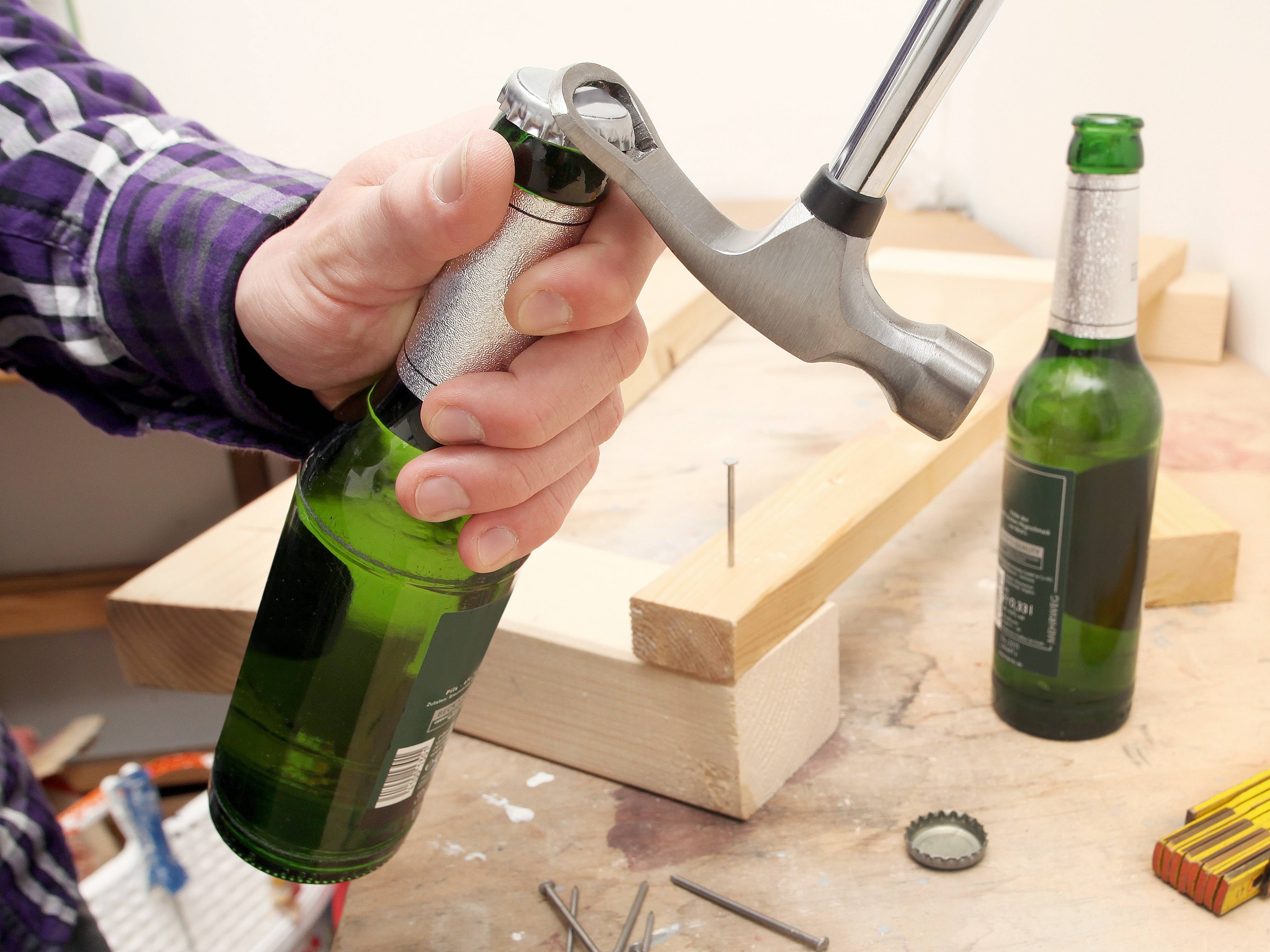 Eine Person benutzt einen Klauhammer, um eine grüne Glasflasche auf einer hölzernen Werkbank zu öffnen. Ein Flaschenöffner und Werkzeuge sind in der Nähe.