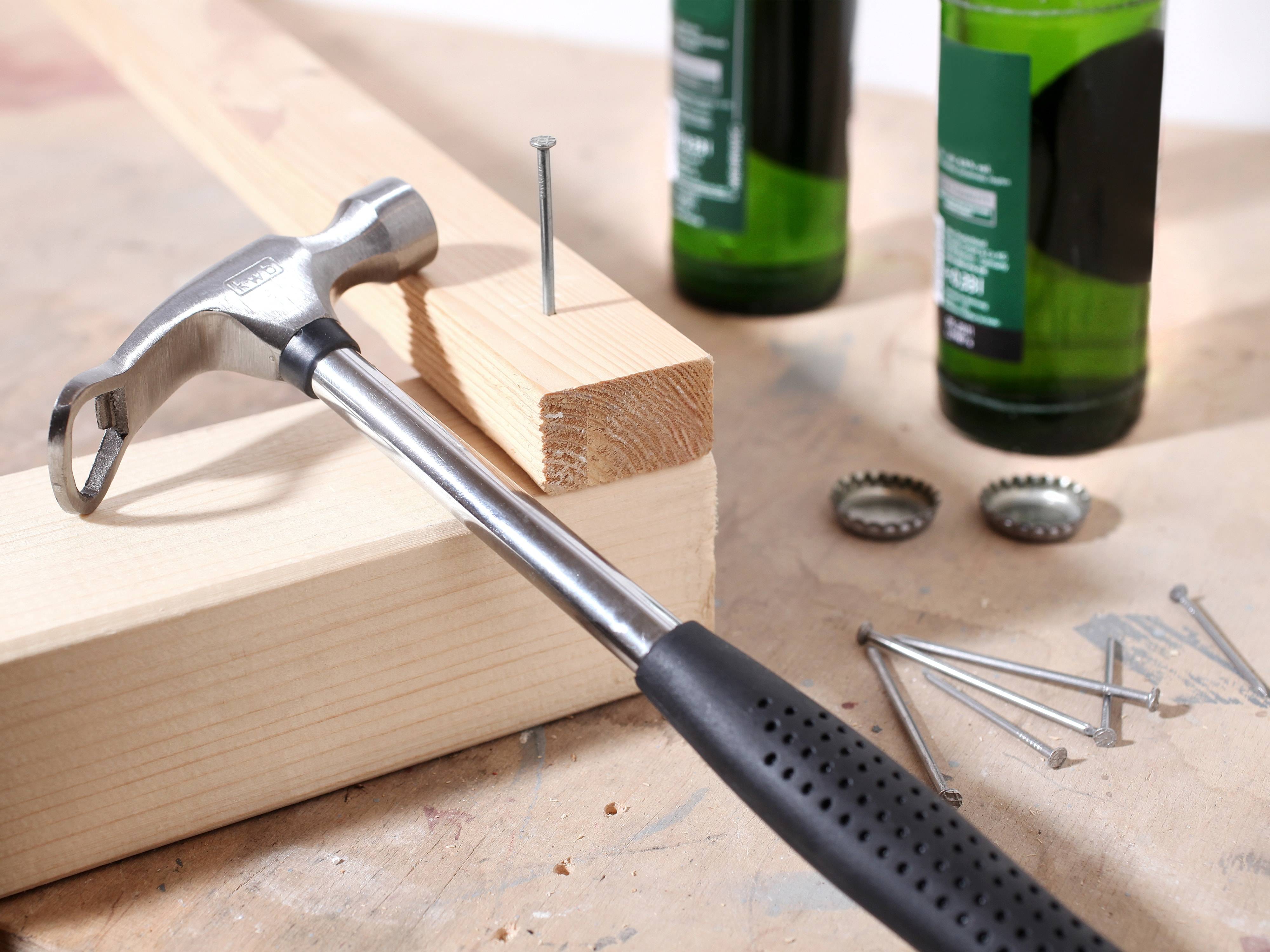 Ein Hammer treibt einen Nagel in einen Holzbalken auf einer Werkbank. Im Hintergrund befinden sich zwei Flaschen und Verschlüsse.