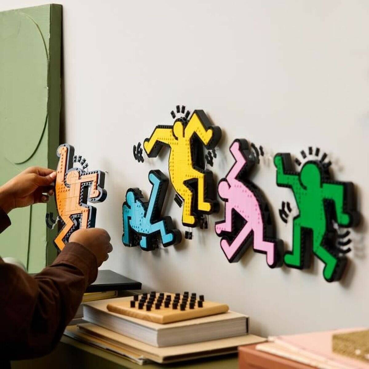Hände, die eine kleine orangefarbene Stiftfigur auf eine Wandanzeige mit bunten Stiftmenschen über gestapelten Büchern und einem Brettspiel platzieren.