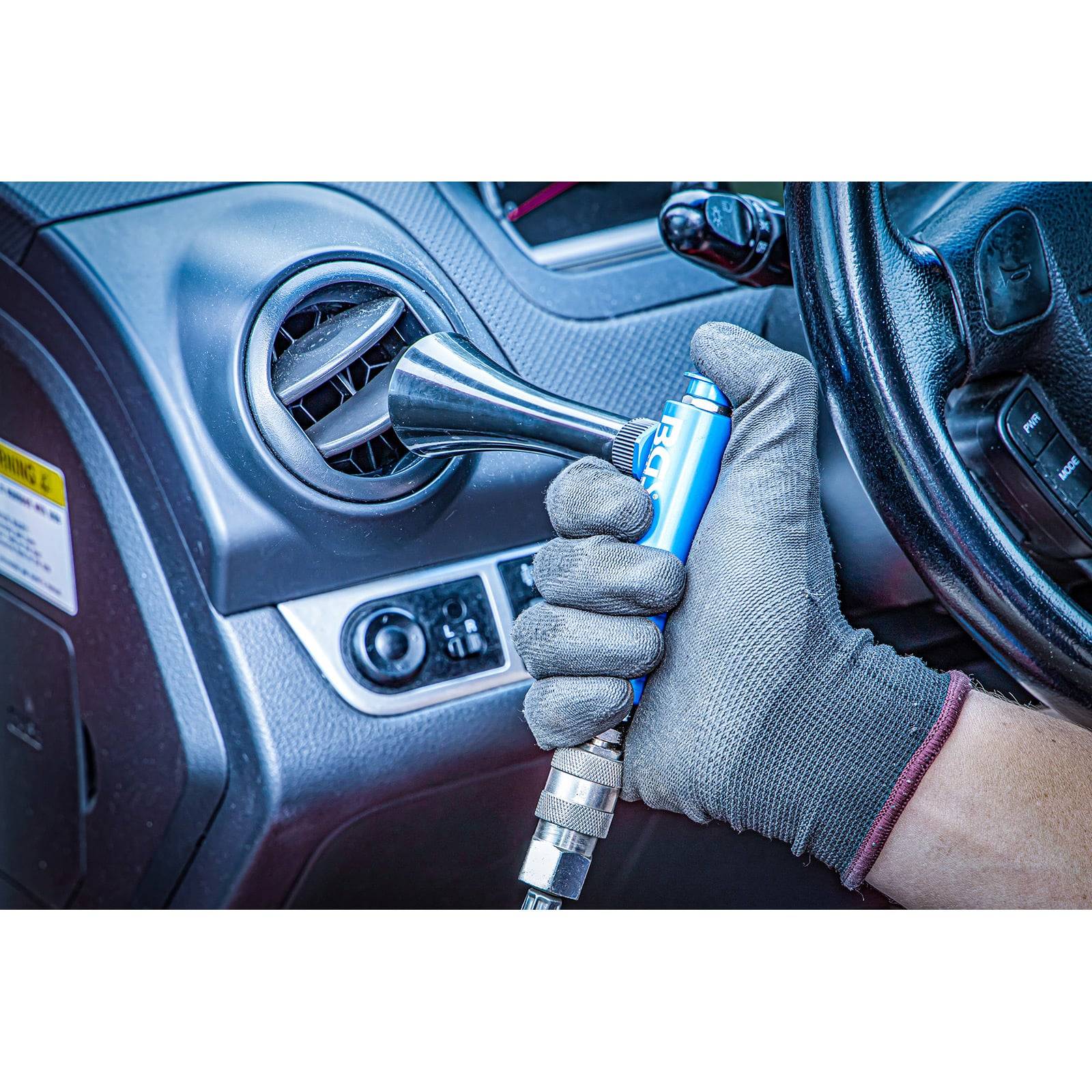 Eine Person verwendet einen tragbaren Luftbläser, um die Luftöffnung eines Autos zu reinigen und trägt dabei einen Handschuh zum Schutz. Das Armaturenbrett und das Lenkrad sind sichtbar.