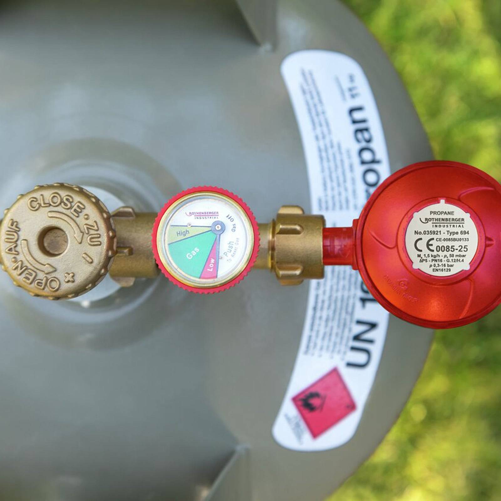 Ein Propangastank mit angeschlossenem Manometer, das einen niedrigen Füllstand anzeigt. Warnschild an der Seite. Helles Außenlicht.