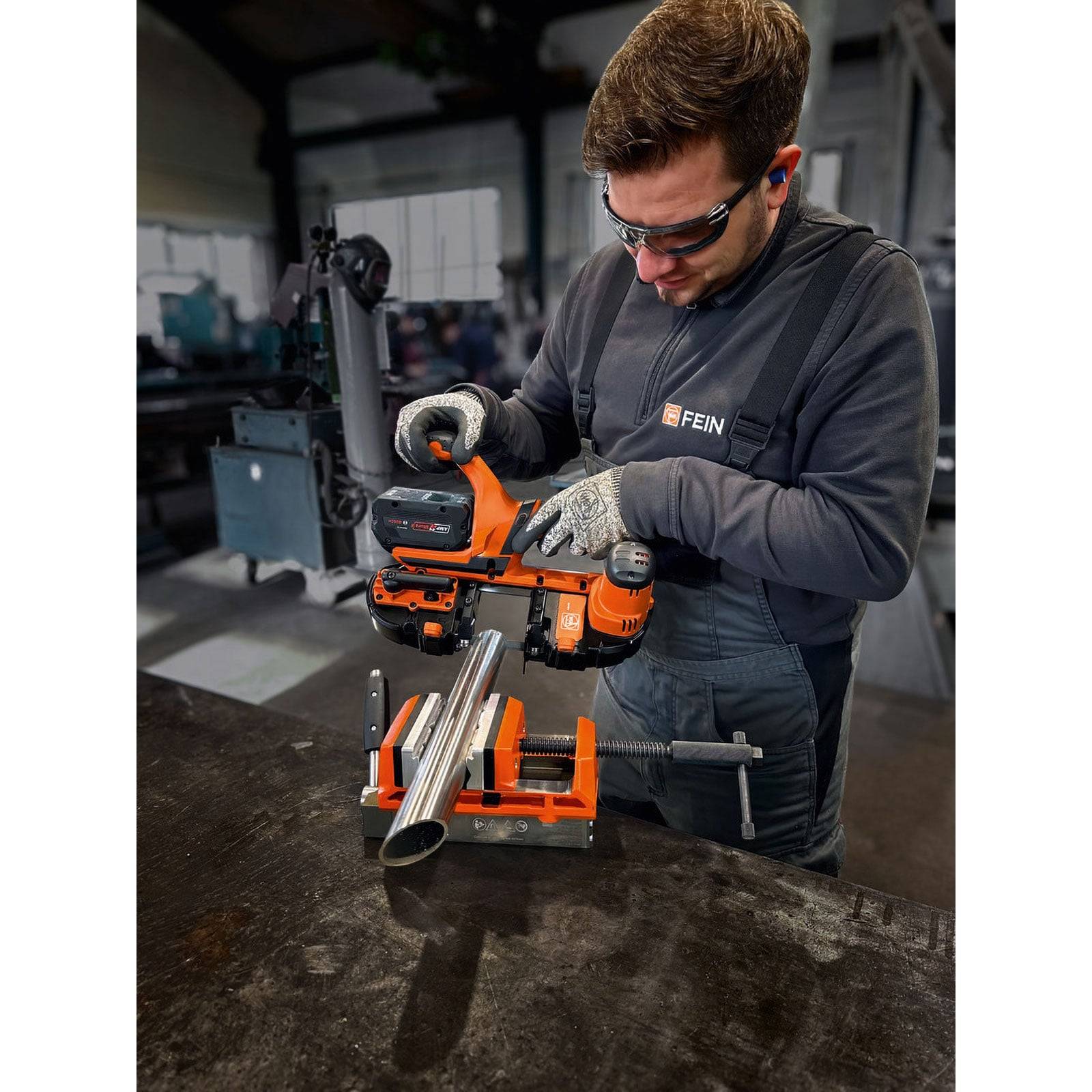 Eine Person in Sicherheitsausrüstung bedient ein orangefarbenes Leistungswerkzeug, um ein Metallrohr auf einer Werkbank in einer industriellen Werkstattumgebung zu schneiden.