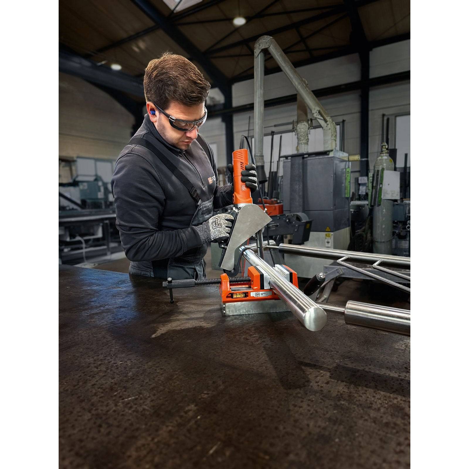 Eine Person in einem Workshop trägt Schutzausrüstung und bedient ein Elektrowerkzeug, wobei sie eine Metallstange auf einer Werkbank bearbeitet, mit industrieller Ausrüstung im Hintergrund.